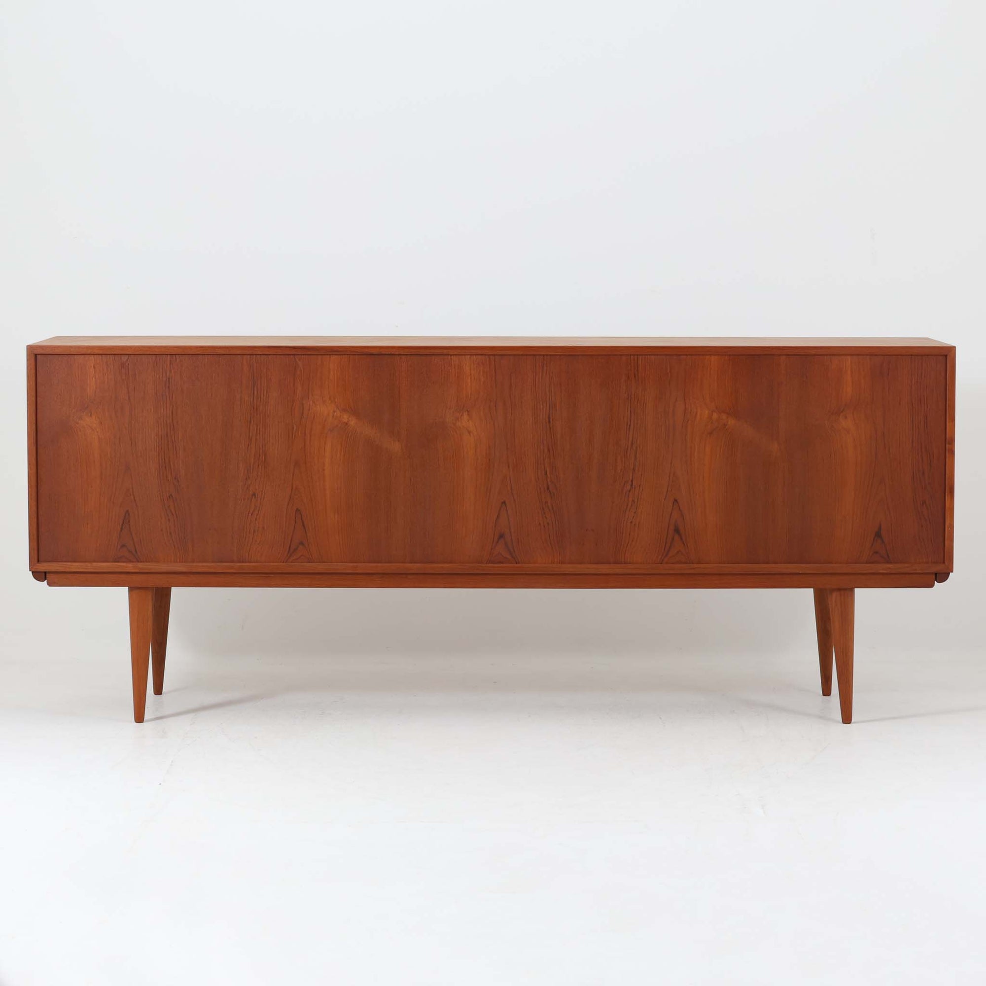 Gunni Omannがデザインし、Omann Jun社によって製造されたModel 18は、洗練されたラインと実用性を兼ね備えた北欧ビンテージの名作サイドボードです。チーク材の美しい木目が前面に広がり、引き出しとスライド扉の組み合わせが高い収納力を実現しています。特に繊細に削り出された取手や先細りの脚部が、軽やかで彫刻的な印象を与え、空間を上質に演出します。幅200cmのサイズ感は、リビングやダイニングの主役として活躍する存在感ある一台です。