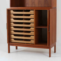 Bjerringbro Savvaerkによるチーク材キャビネットは、上下に分かれた構成と可動棚を備えた収納力の高さが魅力です。上段は3段のシェルフ、下段は珍しいポケットドア構造の扉内にA4サイズの書類が収納可能な引き出し型トレイが8段並び、書類や小物の整理に適しています。直線的で端正なフォルムと、チーク材の木目が美しく調和した佇まいは、北欧ビンテージらしい実用性と美しさを兼ね備えた逸品です。