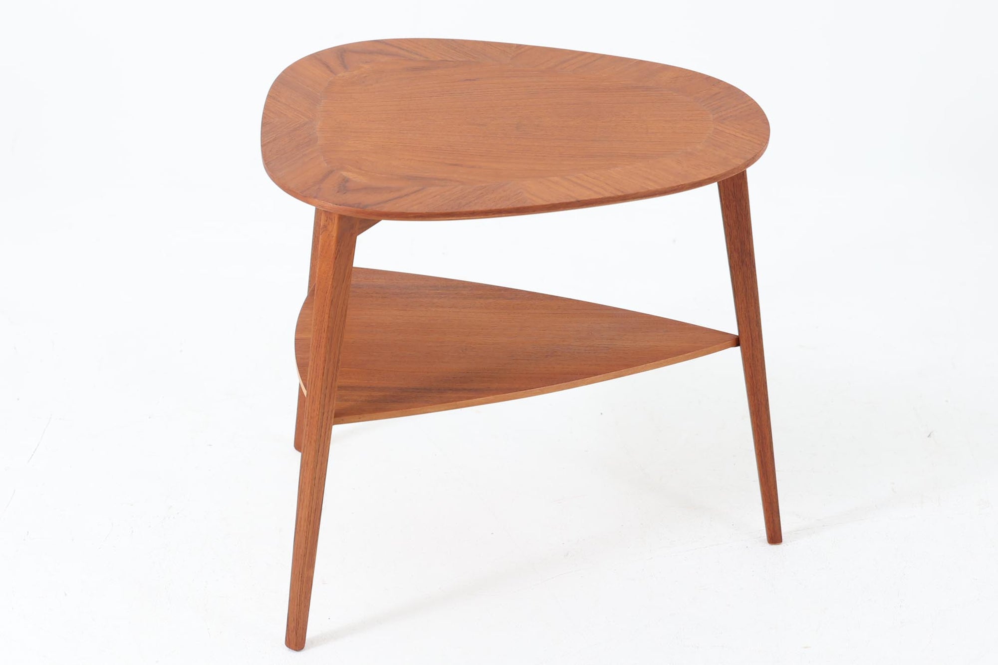 Holger Georg Jensen（ホルガー・ジョージ・イェンセン）によってデザインされ、Kubus moblerから製造されたデンマーク製ビンテージのサイドテーブルです。有機的な曲線を描く天板と、それを支える3本脚の構造が美しいバランスを保っています。天板下には同形状の棚板が設けられており、雑誌や小物の収納にも便利です。素材には艶やかなチーク材が用いられ、天板の木目の美しさも際立っています。北欧モダンを感じさせる上品な佇まいで、ソファサイドやベッドサイドにおすすめの一台です。