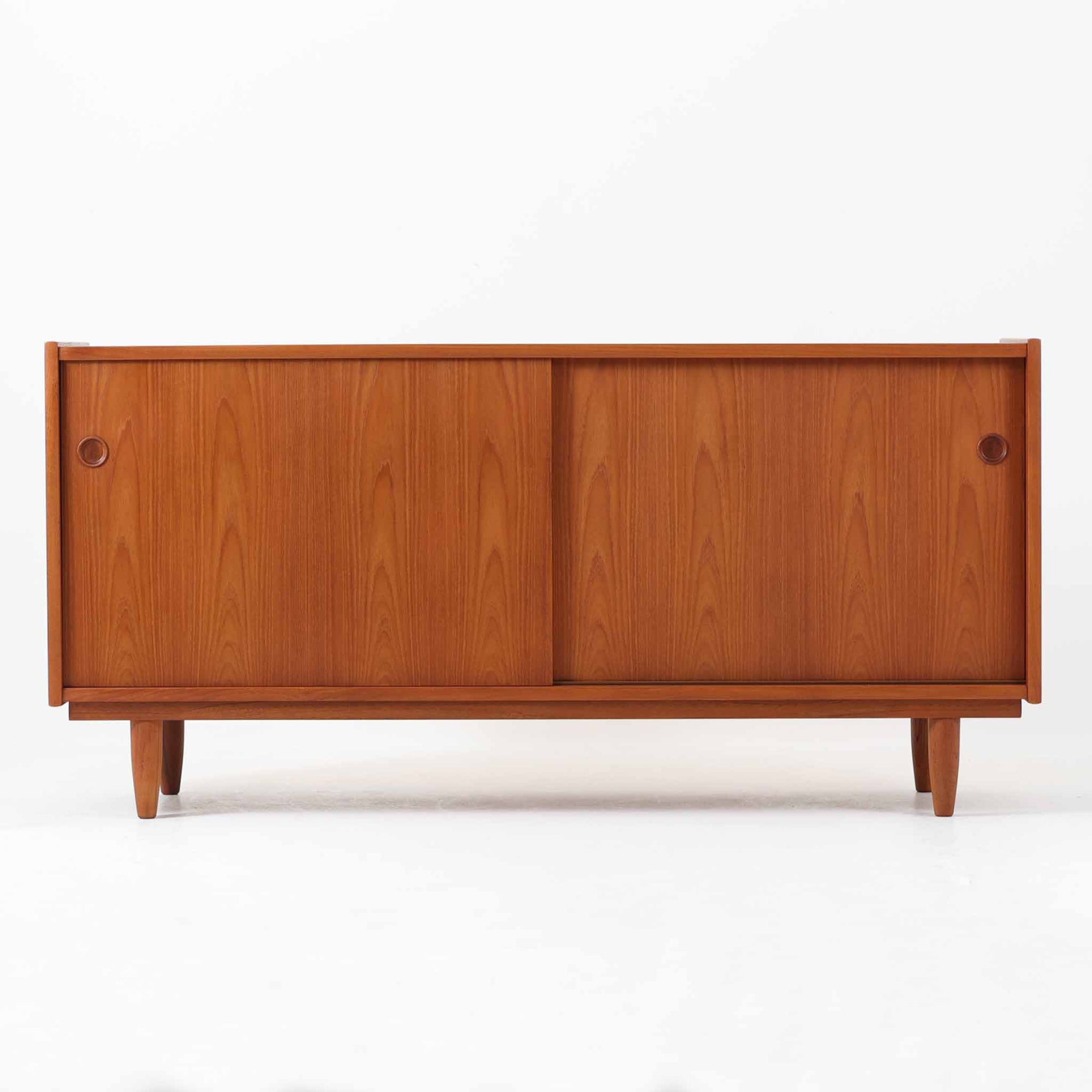 チーク材の美しい木目と滑らかな質感が際立つこちらのローボードは、収納力とデザイン性を兼ね備えた北欧ビンテージ家具です。丸い指掛けが施された引き戸を開けると、内部には仕切り棚やトレイ付きの収納スペースがあり、食器類や小物の整理に最適です。脚部は丸脚で軽やかな印象を与え、リビングのテレビ台やキャビネットとしても活躍する一台です。年月を経て深まった風合いが、空間に温かみと落ち着きをもたらします。