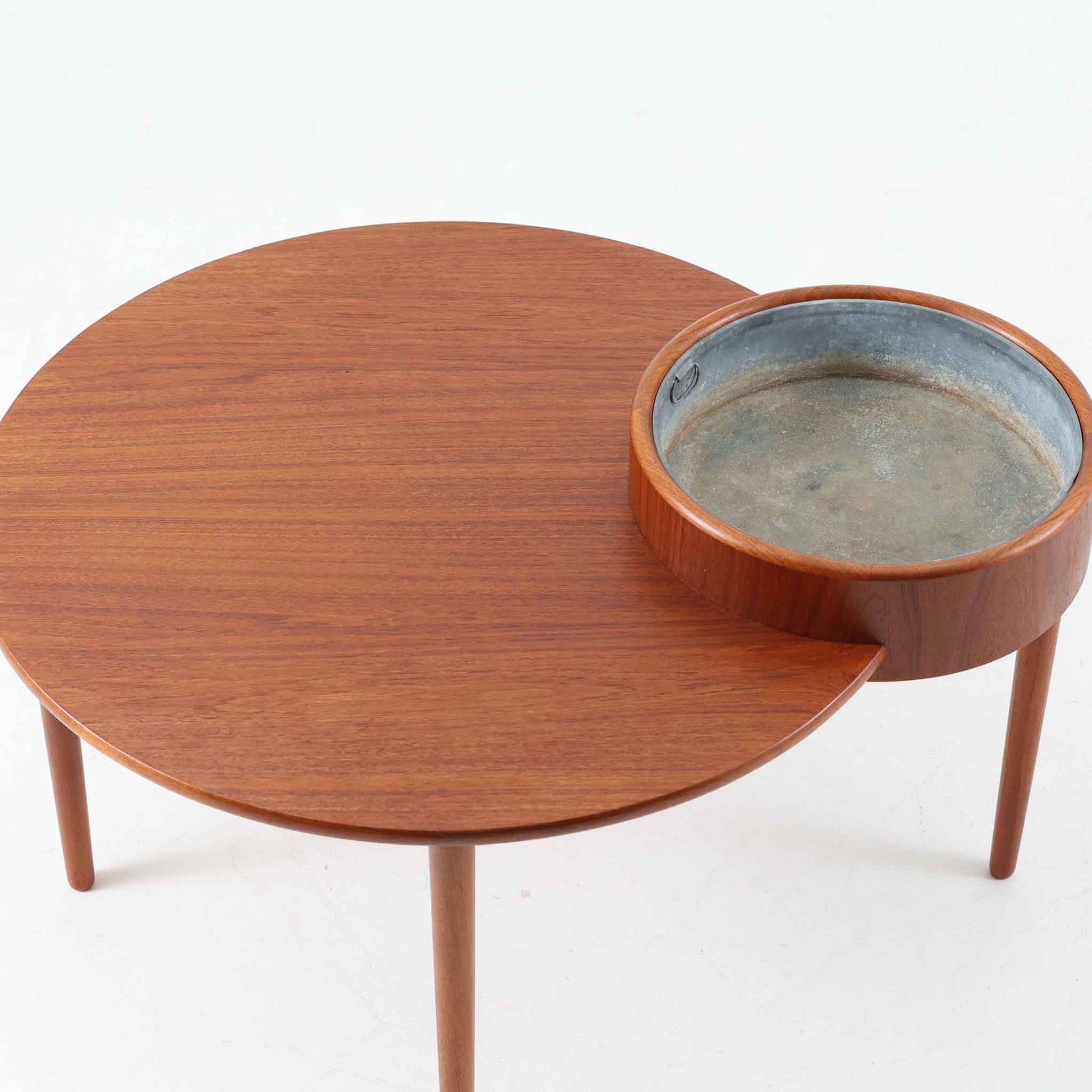 Arne Wahl Iversenがデザインし、Vinde Mobelfabrik社から製造されたユニークなプランター付きセンターテーブルは、チーク材の温かみと実用性を兼ね備えた北欧ビンテージの魅力あふれる一台です。天板の一部にメタル製の植栽スペースが組み込まれており、観葉植物や小物ディスプレイを楽しめる機能的な構造が特徴です。ラウンドフォルムに、スラリと伸びたテーパードレッグが軽やかさを加え、リビング空間に個性と洗練をもたらします。