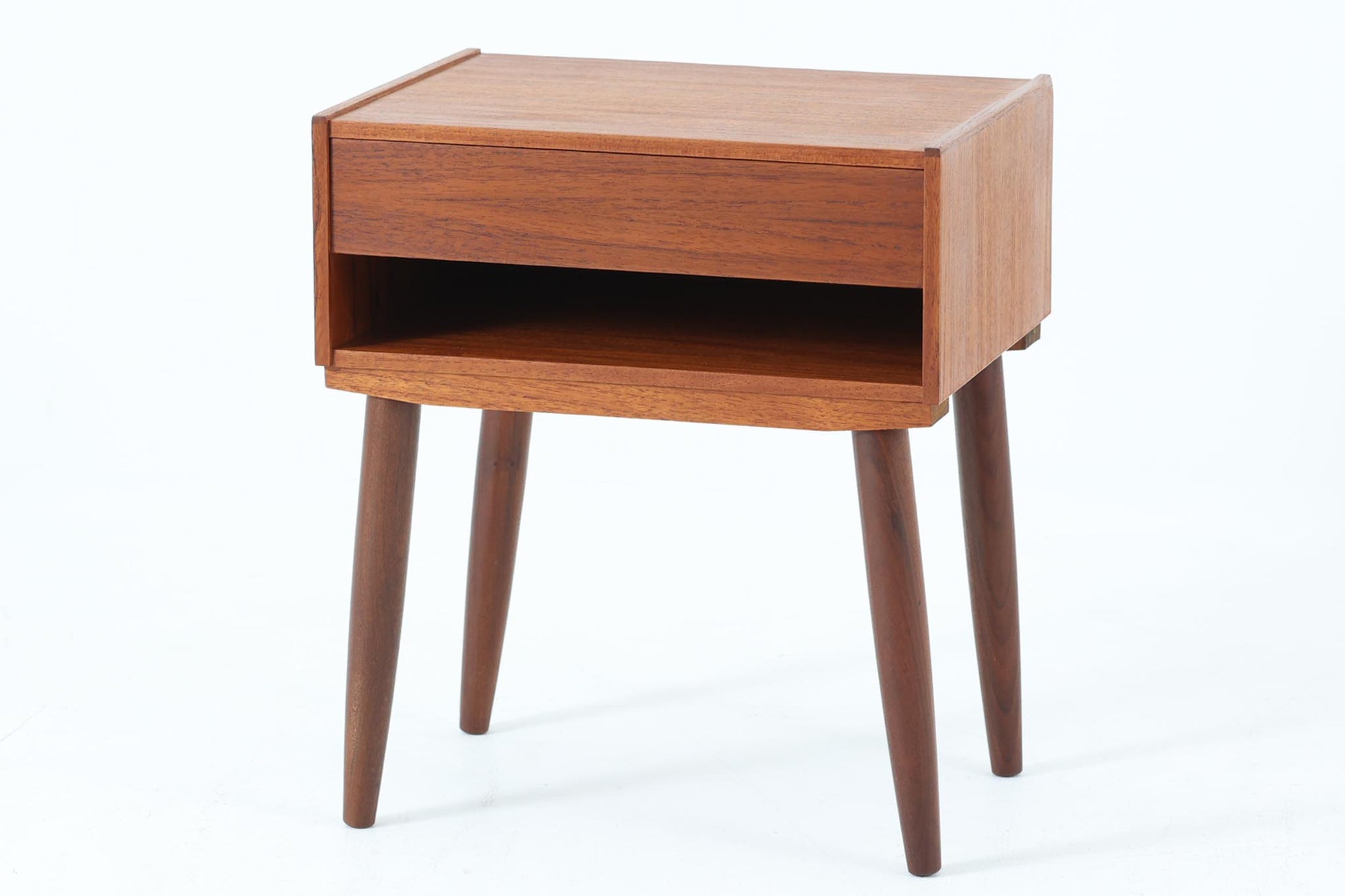 デンマークで製造されたヴィンテージのサイドチェスト。温もりのあるチーク材を用いた本体に、先細りの丸脚を組み合わせた軽やかな佇まいが魅力です。引き出し1杯に加え、下部にはオープンスペースを備えており、リモコンや文庫本などをすぐに取り出せる実用的な構造。天板や側面にはすっきりとした面取りが施され、シンプルなデザインの中に丁寧な仕事が感じられます。北欧らしいあたたかみと実用性を兼ね備えた一台。ベッドサイドはもちろん、リビングや玄関先など、暮らしのさまざまな場面で活躍します。