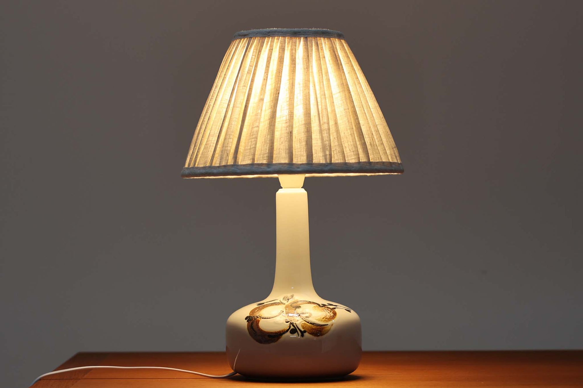 デンマークRoyal Copenhagen製、Ellen malmerデザインによるテーブルランプ。1969〜1974年に製造されたFajanceシリーズの一点で、花を思わせる有機的なモチーフが温かみのある印象を与えます。艶のある白地に手描きで施された装飾が映え、コンパクトながらも空間のアクセントになる存在感があります。シェードは新品のリネン製です。