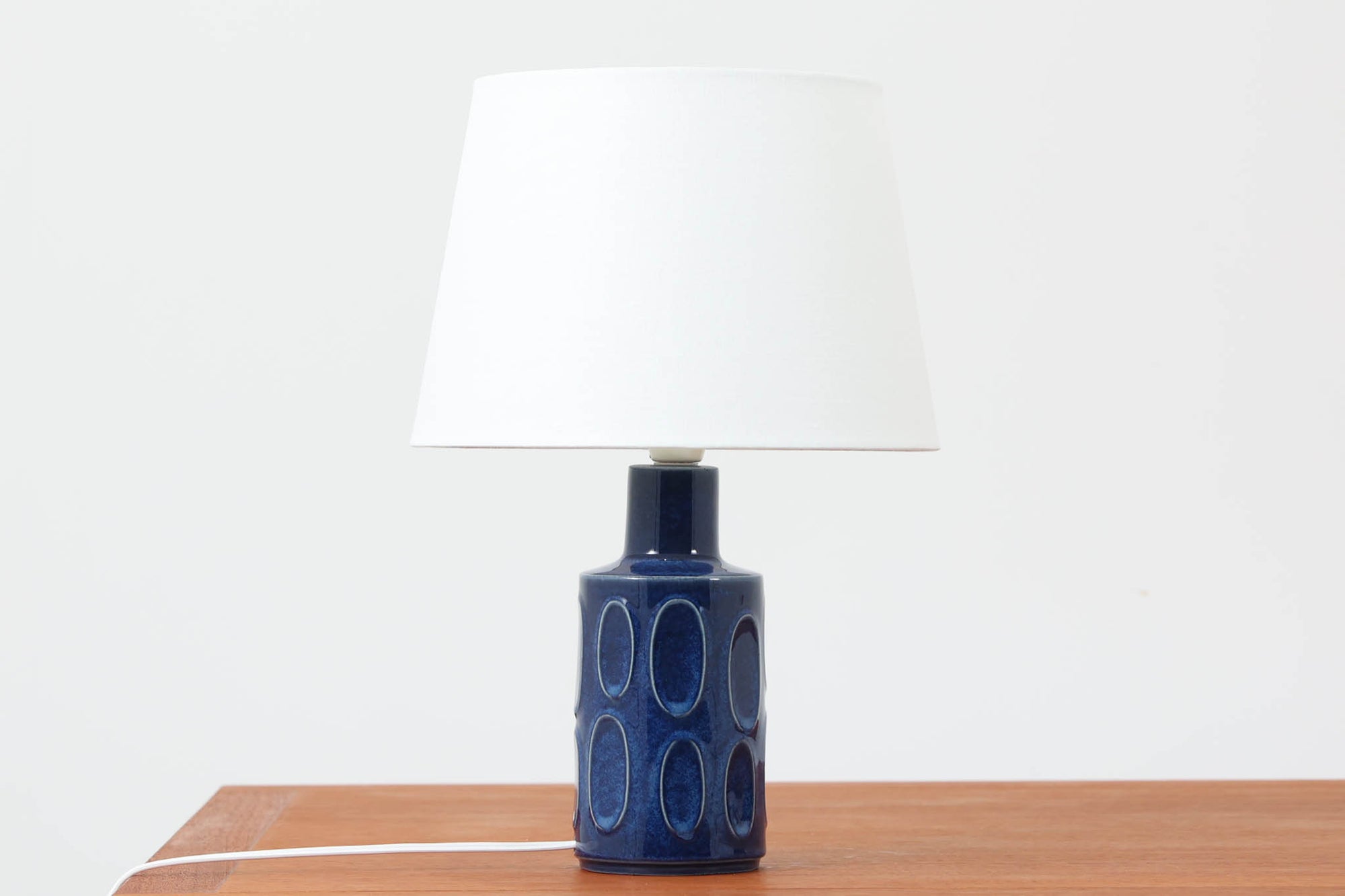 デンマークの陶器ブランドDesiree（デシレ）によって制作されたヴィンテージのテーブルランプ。艶やかな深いブルーの釉薬に、楕円形のレリーフがリズミカルに並ぶ印象的なデザインが魅力です。フォルムはシンプルながらも彫刻的で、北欧陶器ならではの温かみと落ち着きを感じさせます。ベース底部には「Desiree Stentoj – Denmark」の刻印と品番「2010」が施されています。シェードには新品のリネン製シェードを合わせ、光をやわらかく拡散。ベッドサイドやリビングのアクセントとして、空間に静かな存在感を添える一灯です。