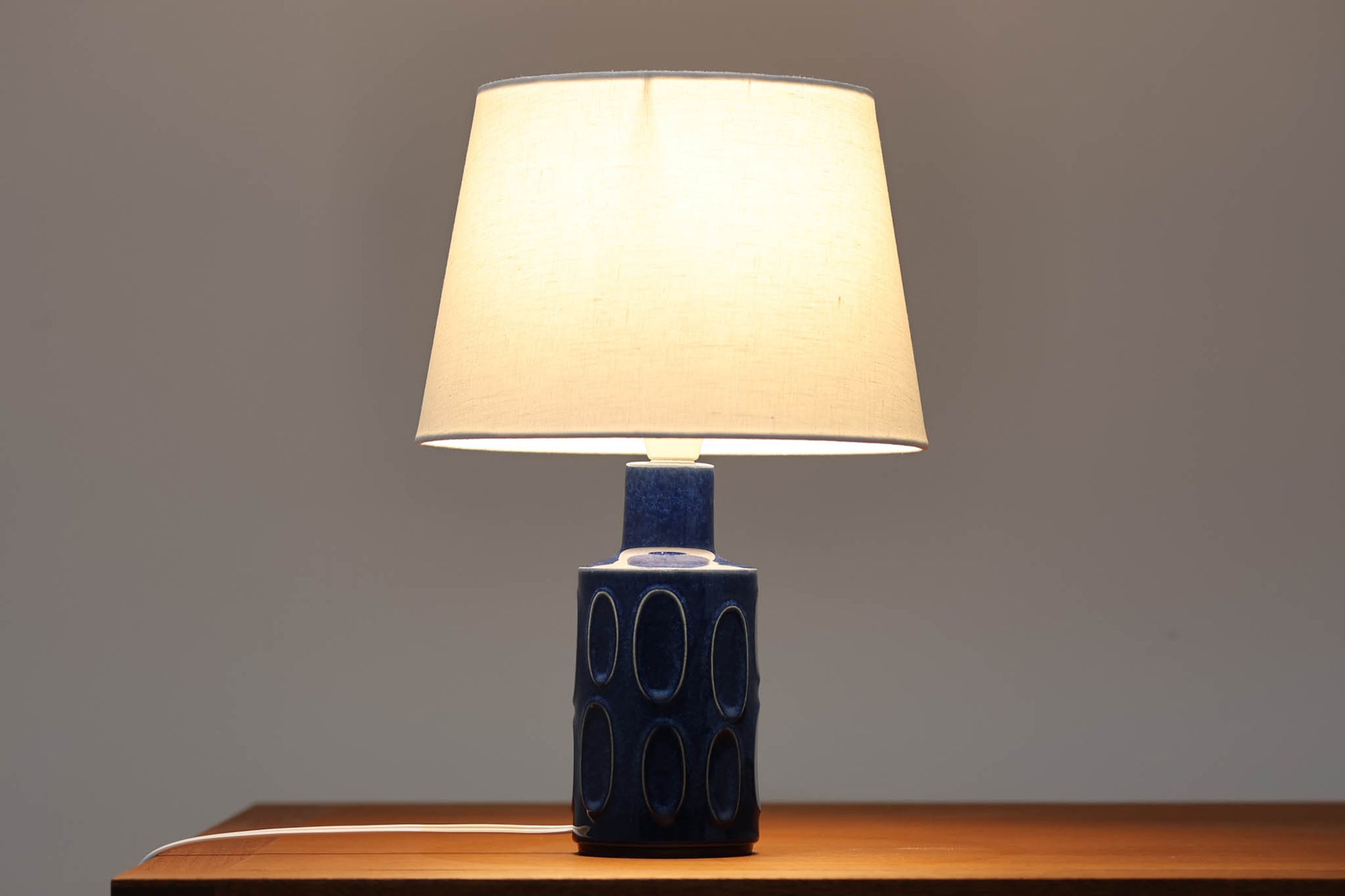 デンマークの陶器ブランドDesiree（デシレ）によって制作されたヴィンテージのテーブルランプ。艶やかな深いブルーの釉薬に、楕円形のレリーフがリズミカルに並ぶ印象的なデザインが魅力です。フォルムはシンプルながらも彫刻的で、北欧陶器ならではの温かみと落ち着きを感じさせます。ベース底部には「Desiree Stentoj – Denmark」の刻印と品番「2010」が施されています。シェードには新品のリネン製シェードを合わせ、光をやわらかく拡散。ベッドサイドやリビングのアクセントとして、空間に静かな存在感を添える一灯です。