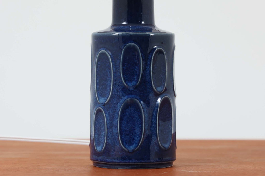 デンマークの陶器ブランドDesiree（デシレ）によって制作されたヴィンテージのテーブルランプ。艶やかな深いブルーの釉薬に、楕円形のレリーフがリズミカルに並ぶ印象的なデザインが魅力です。フォルムはシンプルながらも彫刻的で、北欧陶器ならではの温かみと落ち着きを感じさせます。ベース底部には「Desiree Stentoj – Denmark」の刻印と品番「2010」が施されています。シェードには新品のリネン製シェードを合わせ、光をやわらかく拡散。ベッドサイドやリビングのアクセントとして、空間に静かな存在感を添える一灯です。