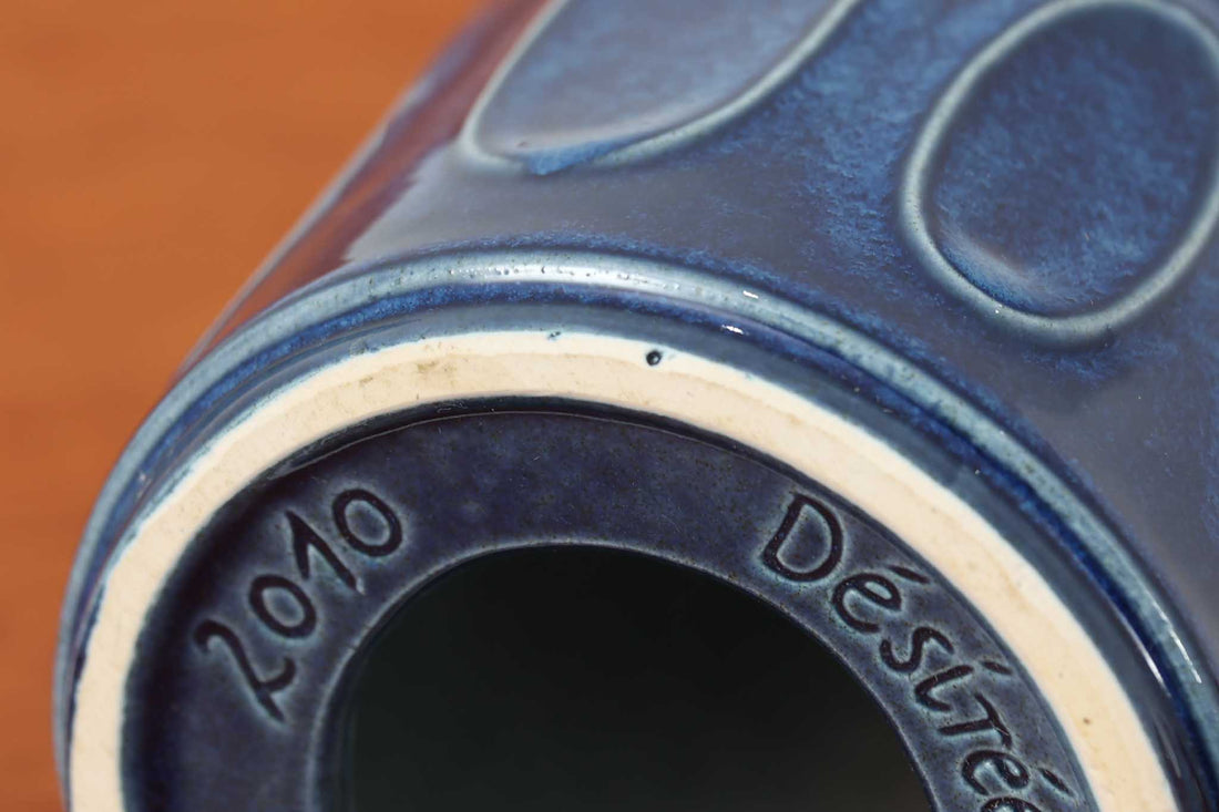 デンマークの陶器ブランドDesiree（デシレ）によって制作されたヴィンテージのテーブルランプ。艶やかな深いブルーの釉薬に、楕円形のレリーフがリズミカルに並ぶ印象的なデザインが魅力です。フォルムはシンプルながらも彫刻的で、北欧陶器ならではの温かみと落ち着きを感じさせます。ベース底部には「Desiree Stentoj – Denmark」の刻印と品番「2010」が施されています。シェードには新品のリネン製シェードを合わせ、光をやわらかく拡散。ベッドサイドやリビングのアクセントとして、空間に静かな存在感を添える一灯です。