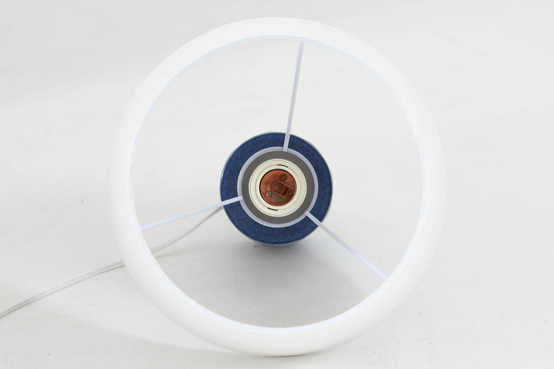 デンマークの陶器ブランドDesiree（デシレ）によって制作されたヴィンテージのテーブルランプ。艶やかな深いブルーの釉薬に、楕円形のレリーフがリズミカルに並ぶ印象的なデザインが魅力です。フォルムはシンプルながらも彫刻的で、北欧陶器ならではの温かみと落ち着きを感じさせます。ベース底部には「Desiree Stentoj – Denmark」の刻印と品番「2010」が施されています。シェードには新品のリネン製シェードを合わせ、光をやわらかく拡散。ベッドサイドやリビングのアクセントとして、空間に静かな存在感を添える一灯です。