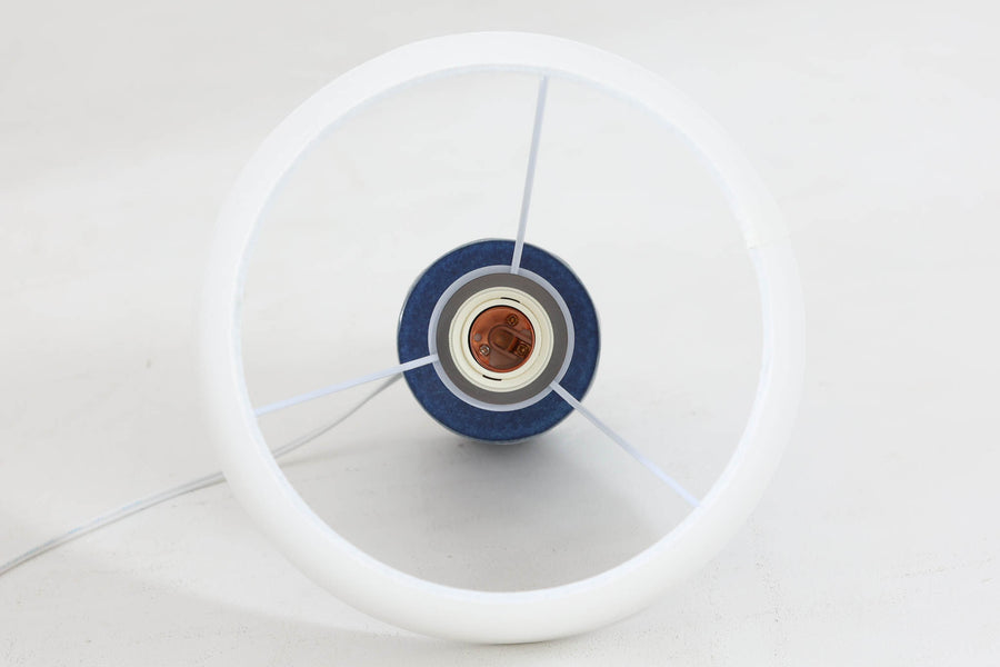 デンマークの陶器ブランドDesiree（デシレ）によって制作されたヴィンテージのテーブルランプ。艶やかな深いブルーの釉薬に、楕円形のレリーフがリズミカルに並ぶ印象的なデザインが魅力です。フォルムはシンプルながらも彫刻的で、北欧陶器ならではの温かみと落ち着きを感じさせます。ベース底部には「Desiree Stentoj – Denmark」の刻印と品番「2010」が施されています。シェードには新品のリネン製シェードを合わせ、光をやわらかく拡散。ベッドサイドやリビングのアクセントとして、空間に静かな存在感を添える一灯です。