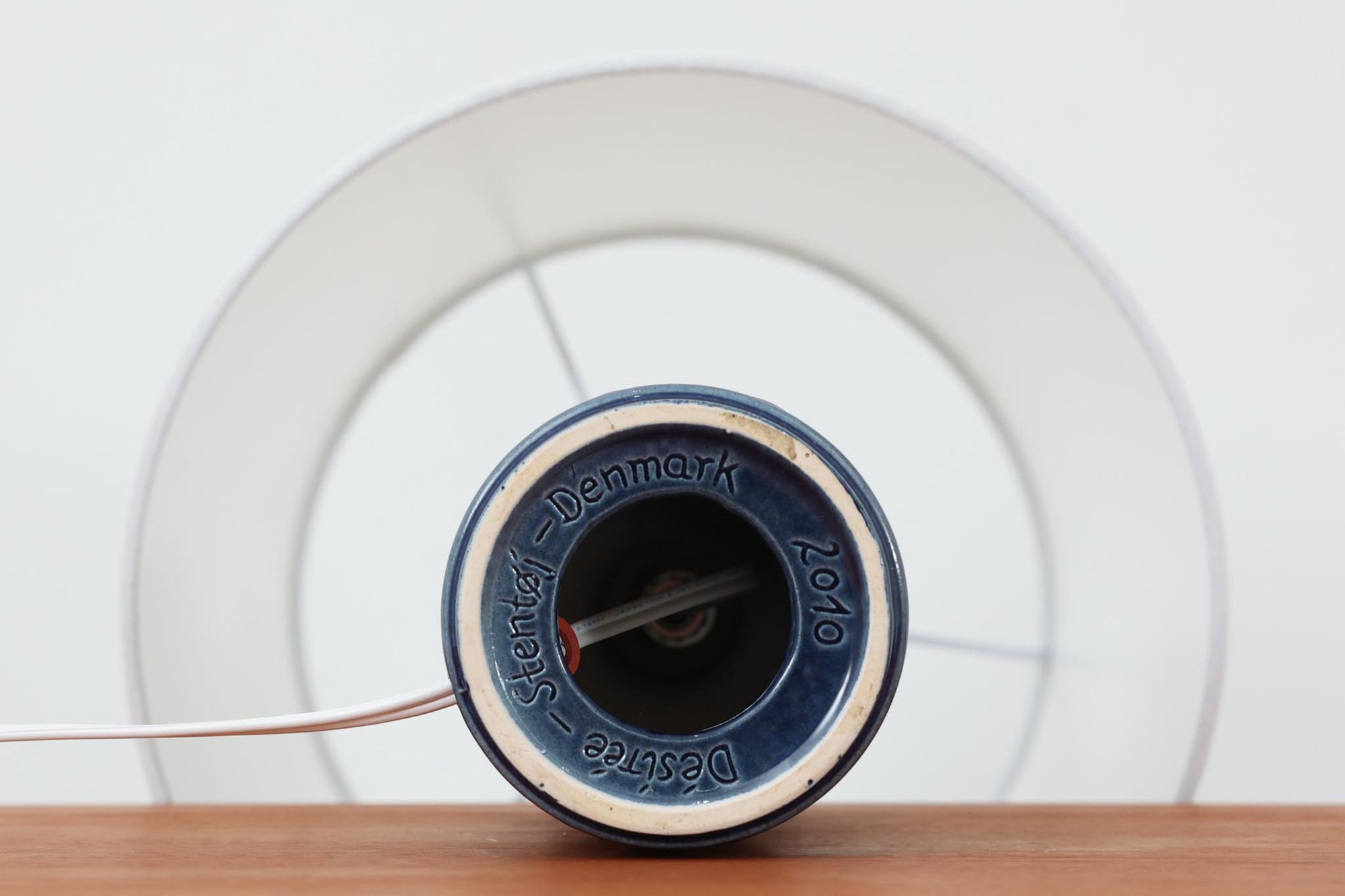 デンマークの陶器ブランドDesiree（デシレ）によって制作されたヴィンテージのテーブルランプ。艶やかな深いブルーの釉薬に、楕円形のレリーフがリズミカルに並ぶ印象的なデザインが魅力です。フォルムはシンプルながらも彫刻的で、北欧陶器ならではの温かみと落ち着きを感じさせます。ベース底部には「Desiree Stentoj – Denmark」の刻印と品番「2010」が施されています。シェードには新品のリネン製シェードを合わせ、光をやわらかく拡散。ベッドサイドやリビングのアクセントとして、空間に静かな存在感を添える一灯です。