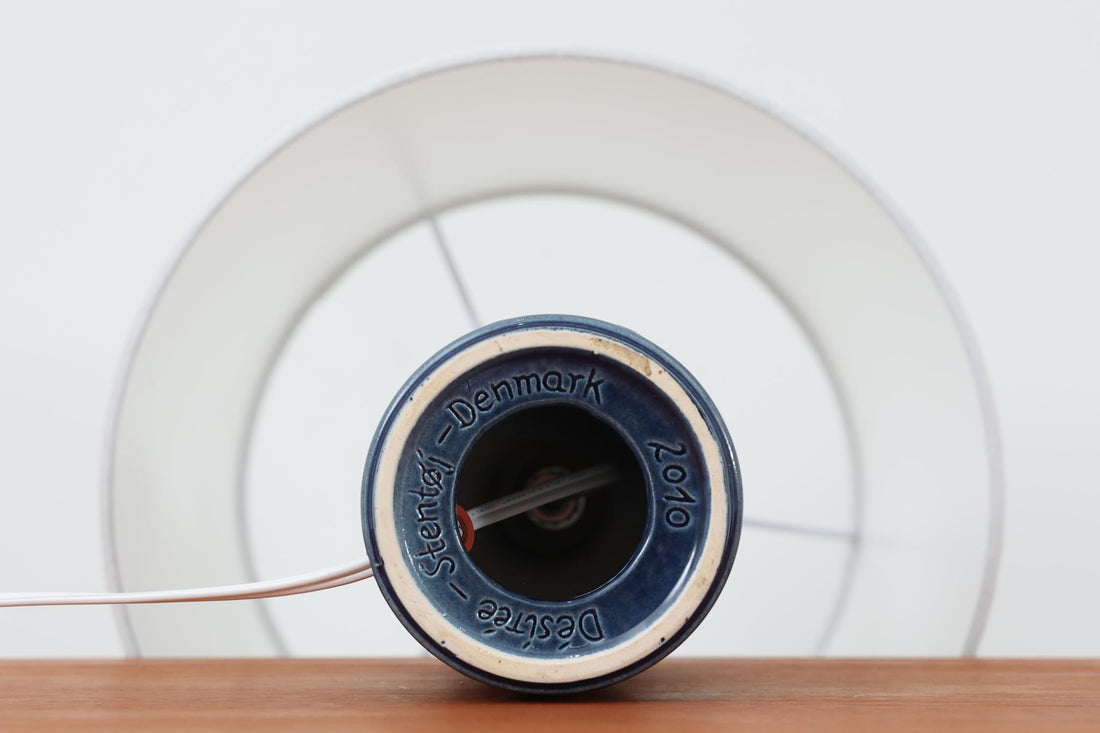 デンマークの陶器ブランドDesiree（デシレ）によって制作されたヴィンテージのテーブルランプ。艶やかな深いブルーの釉薬に、楕円形のレリーフがリズミカルに並ぶ印象的なデザインが魅力です。フォルムはシンプルながらも彫刻的で、北欧陶器ならではの温かみと落ち着きを感じさせます。ベース底部には「Desiree Stentoj – Denmark」の刻印と品番「2010」が施されています。シェードには新品のリネン製シェードを合わせ、光をやわらかく拡散。ベッドサイドやリビングのアクセントとして、空間に静かな存在感を添える一灯です。