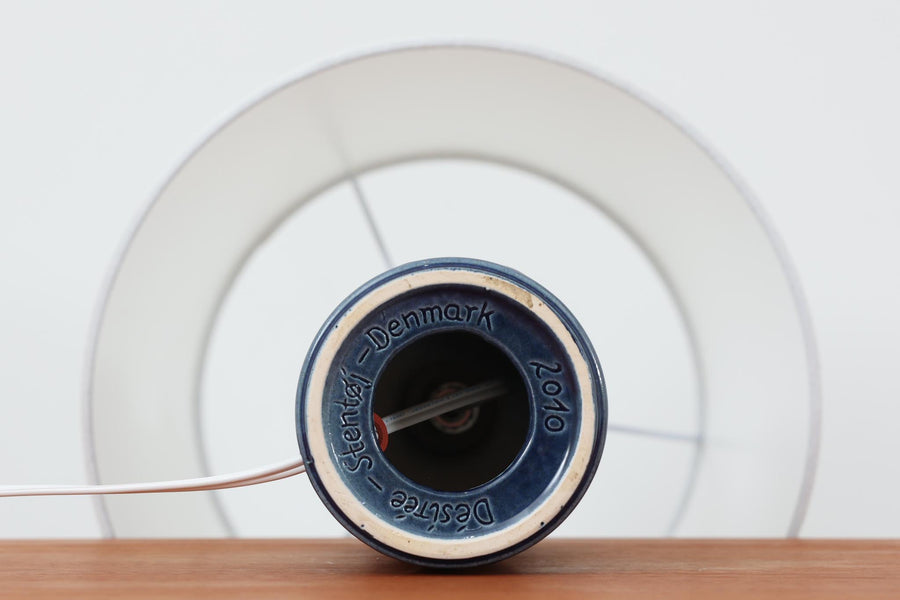 デンマークの陶器ブランドDesiree（デシレ）によって制作されたヴィンテージのテーブルランプ。艶やかな深いブルーの釉薬に、楕円形のレリーフがリズミカルに並ぶ印象的なデザインが魅力です。フォルムはシンプルながらも彫刻的で、北欧陶器ならではの温かみと落ち着きを感じさせます。ベース底部には「Desiree Stentoj – Denmark」の刻印と品番「2010」が施されています。シェードには新品のリネン製シェードを合わせ、光をやわらかく拡散。ベッドサイドやリビングのアクセントとして、空間に静かな存在感を添える一灯です。