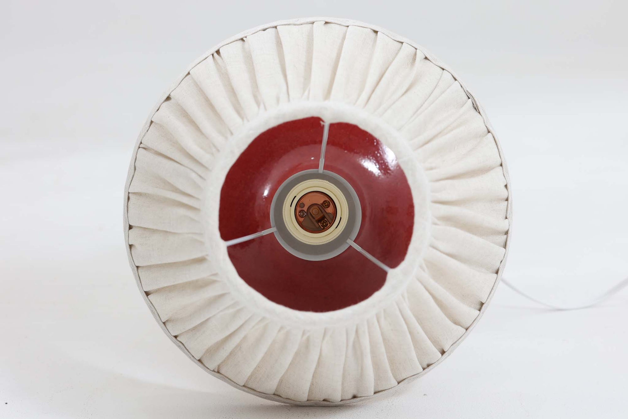 デンマークの照明ブランドLe Klint（レ・クリント）のテーブルランプで、デザイナーはOle Boggild（オーレ・ボギル）。艶やかなレッドの釉薬が美しく、ふっくらと丸みを帯びたフォルムが愛らしくも上品な印象を与えます。シェードはLe Klint社製ではありませんが、新品のリネン製プリーツシェードを合わせており、クラシカルな佇まいと柔らかな光を演出します。北欧らしい温かみと洗練された存在感を持ち、ベッドサイドやリビングのアクセントとして取り入れやすい一灯です。