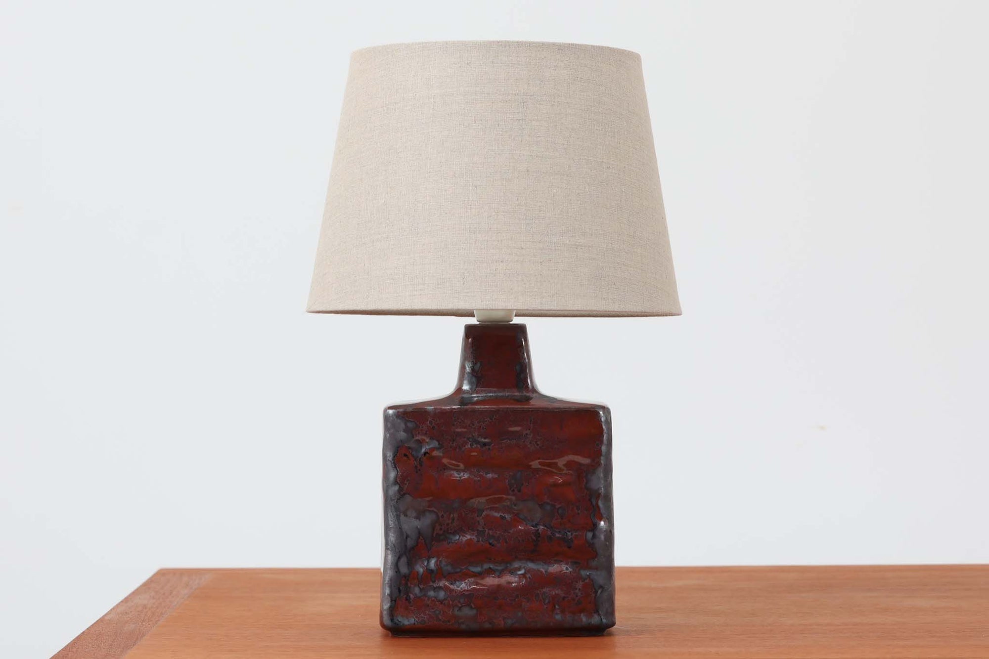 デンマークの陶器ブランドDesiree（デシレ）によって制作されたヴィンテージのテーブルランプ。赤褐色と黒の釉薬が溶け合うように流れ、唯一無二の表情を生み出しています。スクエア型のフォルムに施された厚みのある釉薬の質感は、重厚感と美しさを兼ね備えており、北欧陶器らしい存在感を感じさせます。底部には「Desiree Stentoj – Denmark」の刻印と品番「2071」が見られます。シェードには新品のリネン製を合わせ、やわらかな光が落ち着いた雰囲気を演出します。ヴィンテージらしい個性と静かな力強さを持った一点です。