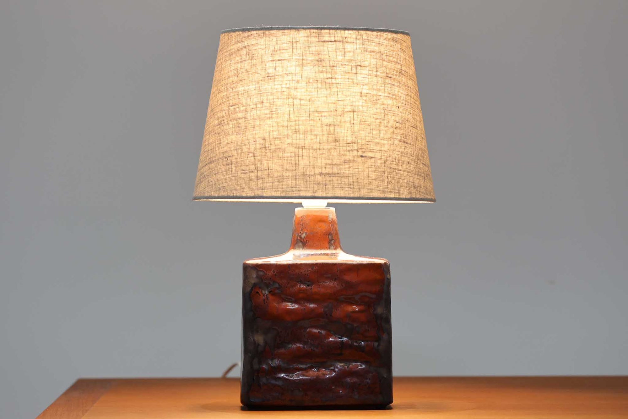 デンマークの陶器ブランドDesiree（デシレ）によって制作されたヴィンテージのテーブルランプ。赤褐色と黒の釉薬が溶け合うように流れ、唯一無二の表情を生み出しています。スクエア型のフォルムに施された厚みのある釉薬の質感は、重厚感と美しさを兼ね備えており、北欧陶器らしい存在感を感じさせます。底部には「Desiree Stentoj – Denmark」の刻印と品番「2071」が見られます。シェードには新品のリネン製を合わせ、やわらかな光が落ち着いた雰囲気を演出します。ヴィンテージらしい個性と静かな力強さを持った一点です。