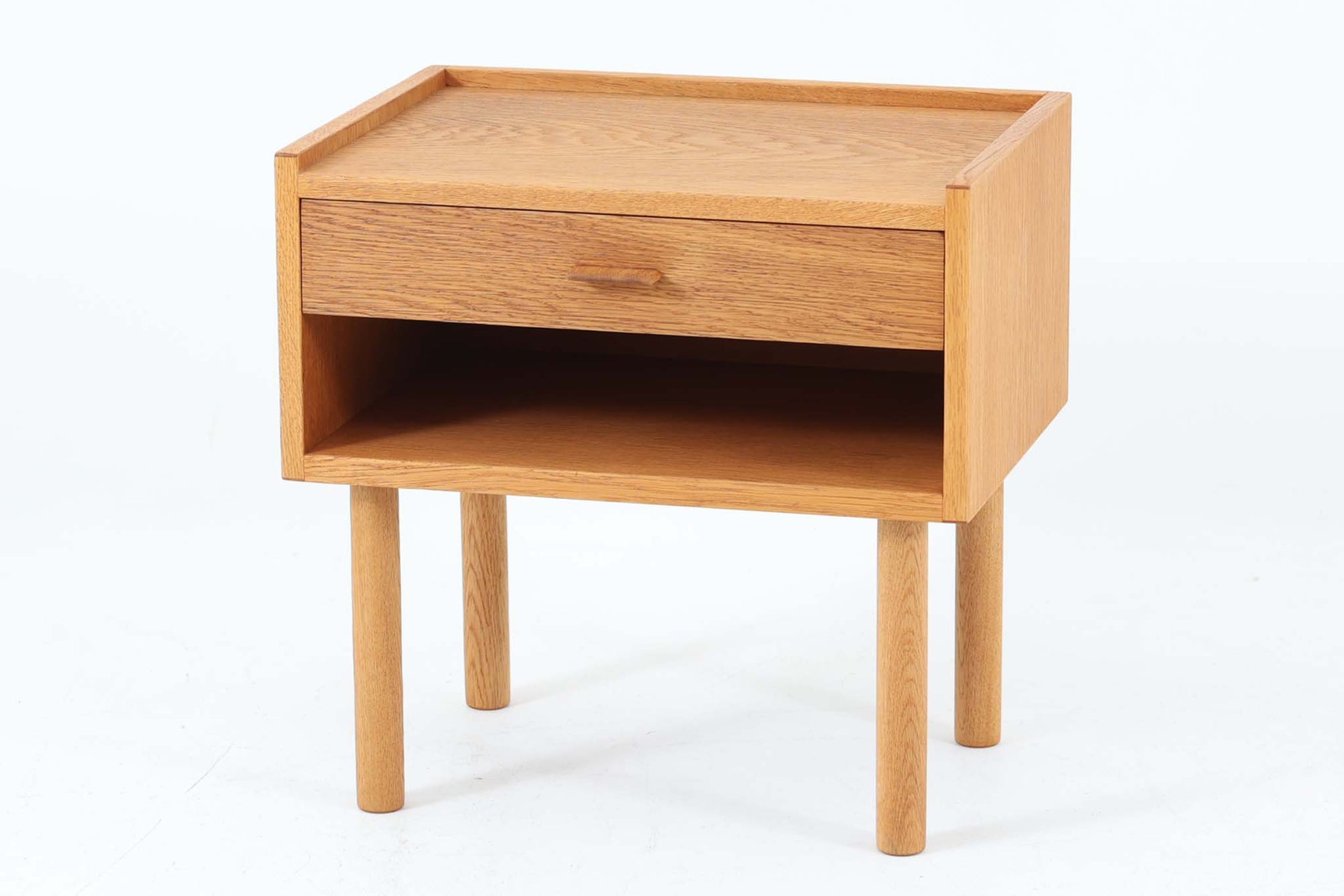 デンマークを代表する家具デザイナー、Hans J. Wegner（ハンス・J・ウェグナー）によってデザインされた、Ry mobler社製のビンテージサイドテーブルです。素材にはあたたかみのあるオーク材が用いられており、北欧らしいシンプルかつ機能的な美しさが感じられます。天板は縁に立ち上がりがあり、物が転がり落ちにくい仕様。引き出しとオープンスペースが組み合わされた実用的な構造で、ベッドサイドやソファ横、玄関周りなど様々なシーンで活躍します。無駄のないデザインと丸脚の柔らかな佇まいが魅力で、ウェグナーデザインならではのバランス感が光る一台です。