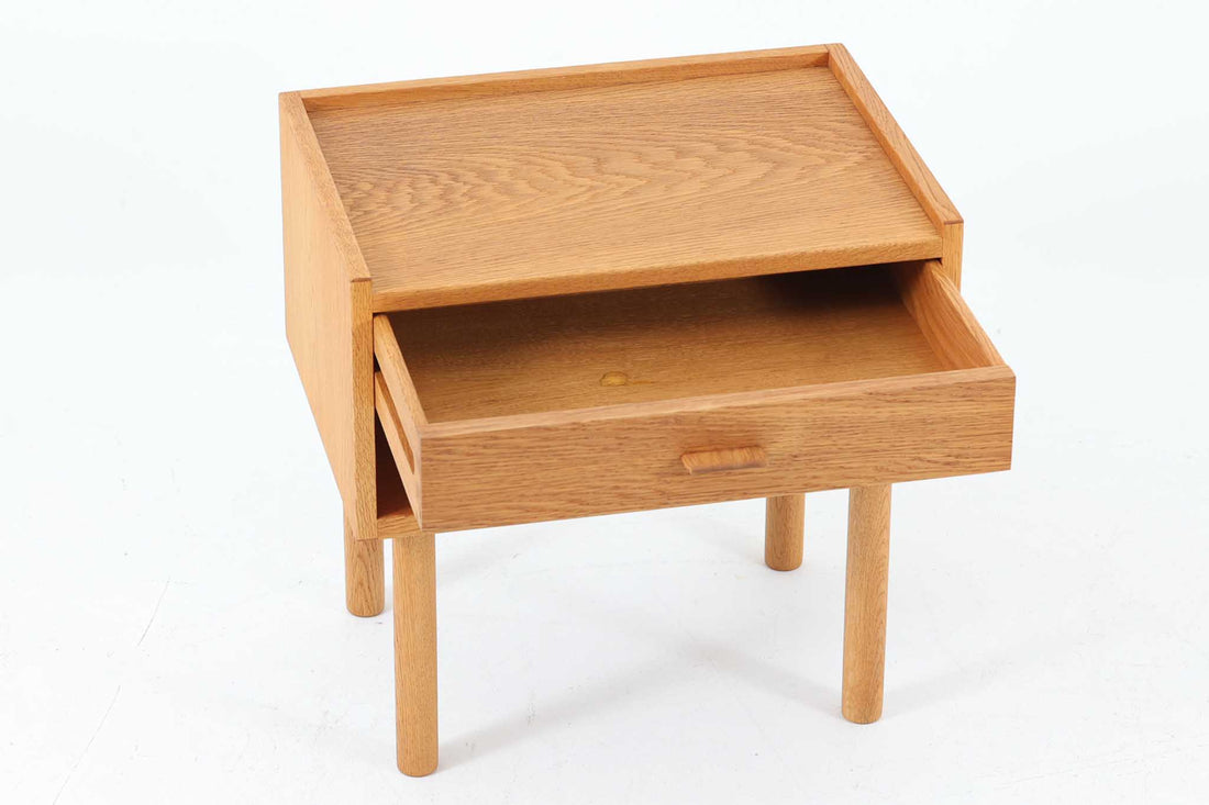 デンマークを代表する家具デザイナー、Hans J. Wegner（ハンス・J・ウェグナー）によってデザインされた、Ry mobler社製のビンテージサイドテーブルです。素材にはあたたかみのあるオーク材が用いられており、北欧らしいシンプルかつ機能的な美しさが感じられます。天板は縁に立ち上がりがあり、物が転がり落ちにくい仕様。引き出しとオープンスペースが組み合わされた実用的な構造で、ベッドサイドやソファ横、玄関周りなど様々なシーンで活躍します。無駄のないデザインと丸脚の柔らかな佇まいが魅力で、ウェグナーデザインならではのバランス感が光る一台です。