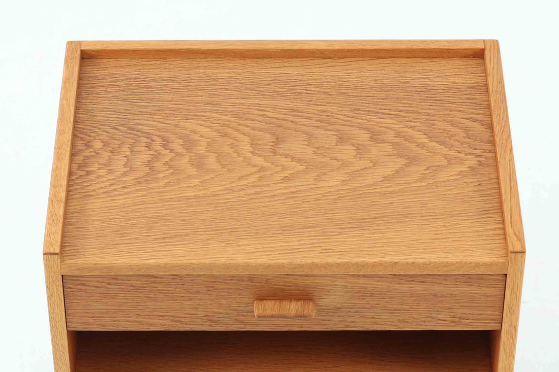 デンマークを代表する家具デザイナー、Hans J. Wegner（ハンス・J・ウェグナー）によってデザインされた、Ry mobler社製のビンテージサイドテーブルです。素材にはあたたかみのあるオーク材が用いられており、北欧らしいシンプルかつ機能的な美しさが感じられます。天板は縁に立ち上がりがあり、物が転がり落ちにくい仕様。引き出しとオープンスペースが組み合わされた実用的な構造で、ベッドサイドやソファ横、玄関周りなど様々なシーンで活躍します。無駄のないデザインと丸脚の柔らかな佇まいが魅力で、ウェグナーデザインならではのバランス感が光る一台です。