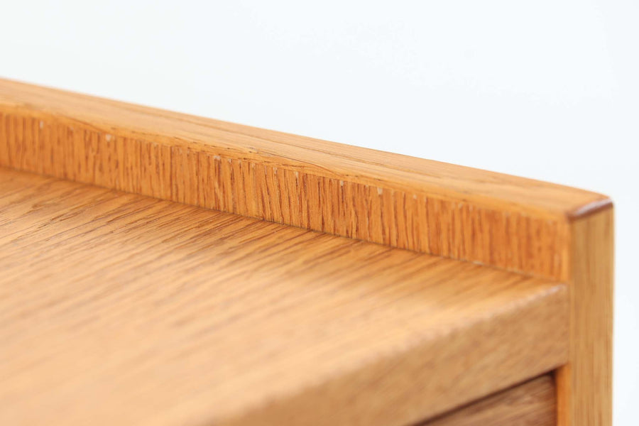 デンマークを代表する家具デザイナー、Hans J. Wegner（ハンス・J・ウェグナー）によってデザインされた、Ry mobler社製のビンテージサイドテーブルです。素材にはあたたかみのあるオーク材が用いられており、北欧らしいシンプルかつ機能的な美しさが感じられます。天板は縁に立ち上がりがあり、物が転がり落ちにくい仕様。引き出しとオープンスペースが組み合わされた実用的な構造で、ベッドサイドやソファ横、玄関周りなど様々なシーンで活躍します。無駄のないデザインと丸脚の柔らかな佇まいが魅力で、ウェグナーデザインならではのバランス感が光る一台です。