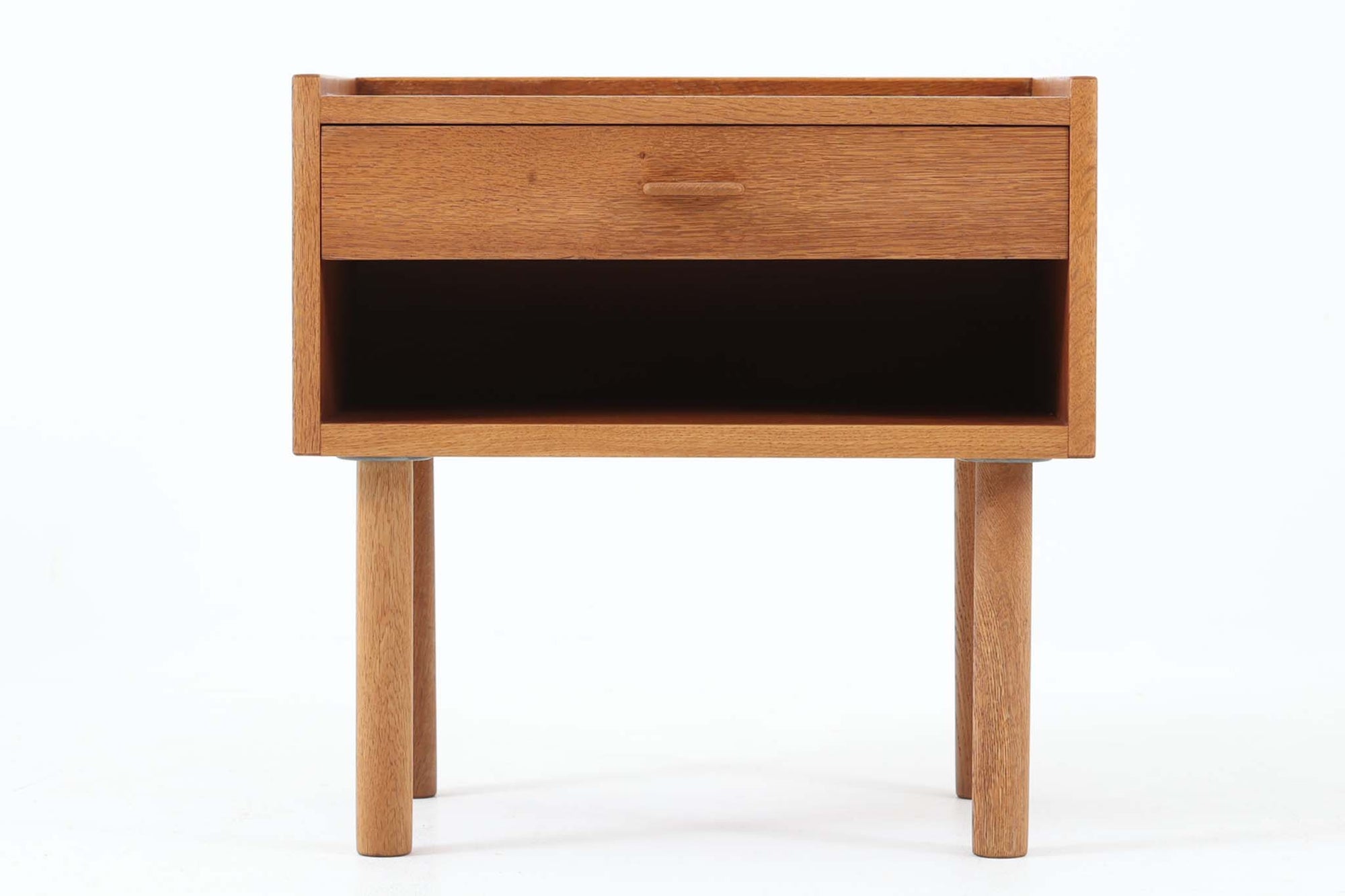 デンマークを代表する家具デザイナー、Hans J. Wegner（ハンス・J・ウェグナー）によってデザインされた、Ry mobler社製のビンテージサイドテーブルです。素材にはあたたかみのあるオーク材が使用されており、北欧らしいシンプルで機能的な美しさが感じられます。天板には縁に立ち上がりが設けられており、小物が転がり落ちにくい実用的な仕様。引き出しとオープンスペースが組み合わされたデザインで、ベッドサイドやソファ横、玄関周りなど様々なシーンで活躍します。無駄のないフォルムと丸脚の柔らかな佇まいが魅力で、ウェグナーデザインならではの美しいバランスが光る一台です。背板には、同モデルとしては珍しくオーク材ではなくmDFと思われる素材が使用されています。また、天板には目立つ傷がございますため、その点を考慮した価格設定とさせていただいております。