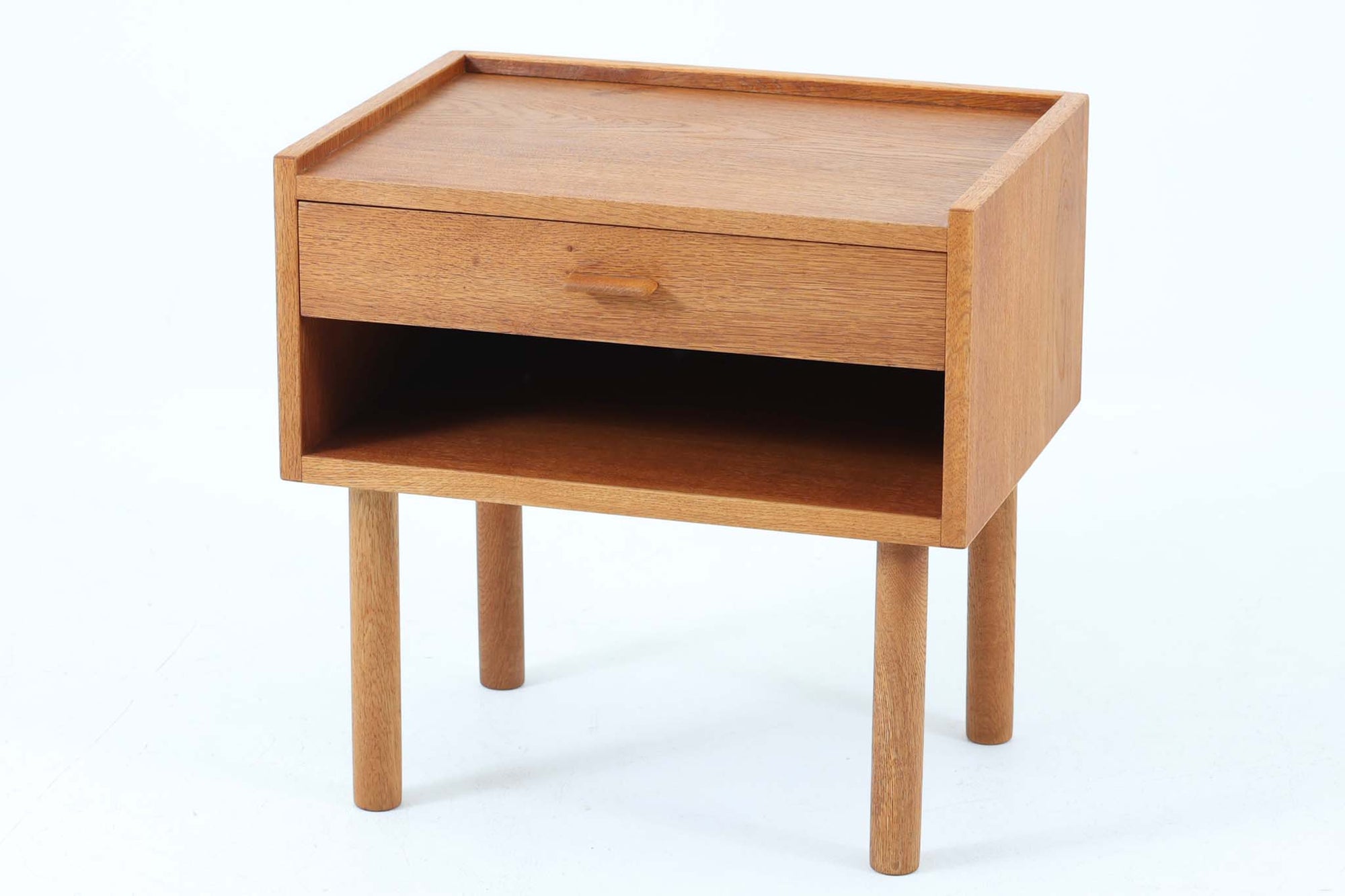 デンマークを代表する家具デザイナー、Hans J. Wegner（ハンス・J・ウェグナー）によってデザインされた、Ry mobler社製のビンテージサイドテーブルです。素材にはあたたかみのあるオーク材が使用されており、北欧らしいシンプルで機能的な美しさが感じられます。天板には縁に立ち上がりが設けられており、小物が転がり落ちにくい実用的な仕様。引き出しとオープンスペースが組み合わされたデザインで、ベッドサイドやソファ横、玄関周りなど様々なシーンで活躍します。無駄のないフォルムと丸脚の柔らかな佇まいが魅力で、ウェグナーデザインならではの美しいバランスが光る一台です。背板には、同モデルとしては珍しくオーク材ではなくmDFと思われる素材が使用されています。また、天板には目立つ傷がございますため、その点を考慮した価格設定とさせていただいております。