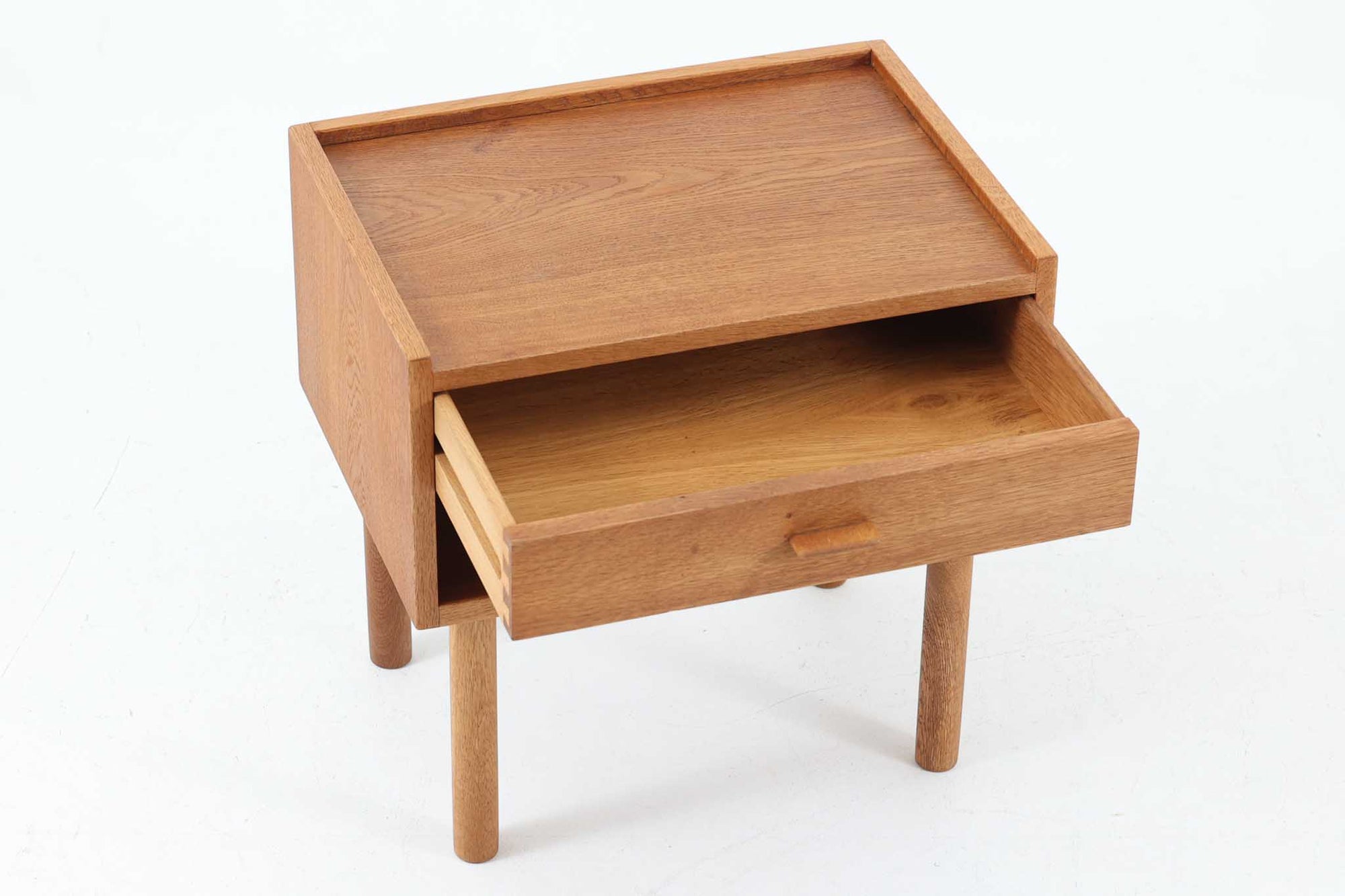 デンマークを代表する家具デザイナー、Hans J. Wegner（ハンス・J・ウェグナー）によってデザインされた、Ry mobler社製のビンテージサイドテーブルです。素材にはあたたかみのあるオーク材が使用されており、北欧らしいシンプルで機能的な美しさが感じられます。天板には縁に立ち上がりが設けられており、小物が転がり落ちにくい実用的な仕様。引き出しとオープンスペースが組み合わされたデザインで、ベッドサイドやソファ横、玄関周りなど様々なシーンで活躍します。無駄のないフォルムと丸脚の柔らかな佇まいが魅力で、ウェグナーデザインならではの美しいバランスが光る一台です。背板には、同モデルとしては珍しくオーク材ではなくmDFと思われる素材が使用されています。また、天板には目立つ傷がございますため、その点を考慮した価格設定とさせていただいております。