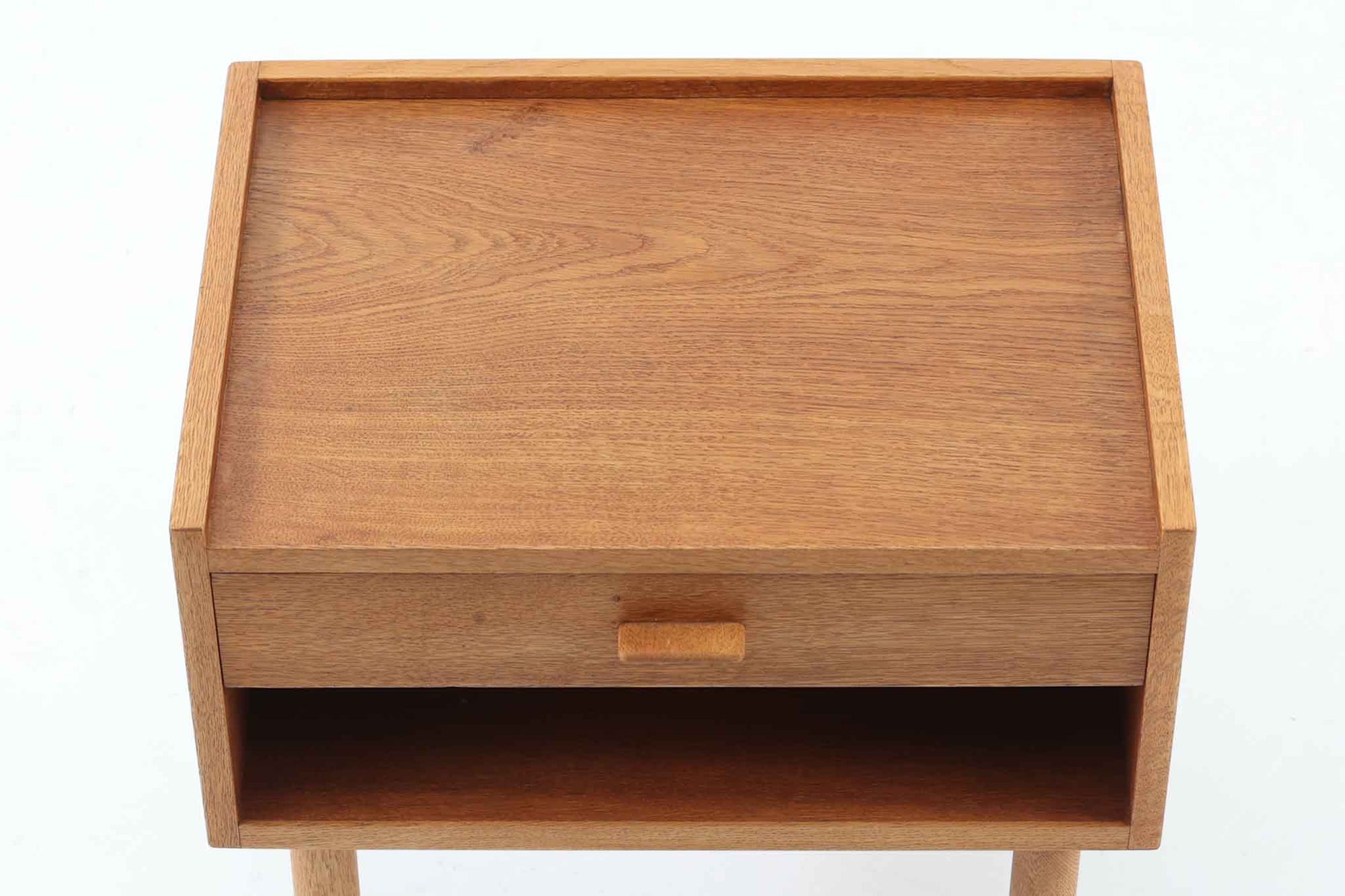 デンマークを代表する家具デザイナー、Hans J. Wegner（ハンス・J・ウェグナー）によってデザインされた、Ry mobler社製のビンテージサイドテーブルです。素材にはあたたかみのあるオーク材が使用されており、北欧らしいシンプルで機能的な美しさが感じられます。天板には縁に立ち上がりが設けられており、小物が転がり落ちにくい実用的な仕様。引き出しとオープンスペースが組み合わされたデザインで、ベッドサイドやソファ横、玄関周りなど様々なシーンで活躍します。無駄のないフォルムと丸脚の柔らかな佇まいが魅力で、ウェグナーデザインならではの美しいバランスが光る一台です。背板には、同モデルとしては珍しくオーク材ではなくmDFと思われる素材が使用されています。また、天板には目立つ傷がございますため、その点を考慮した価格設定とさせていただいております。