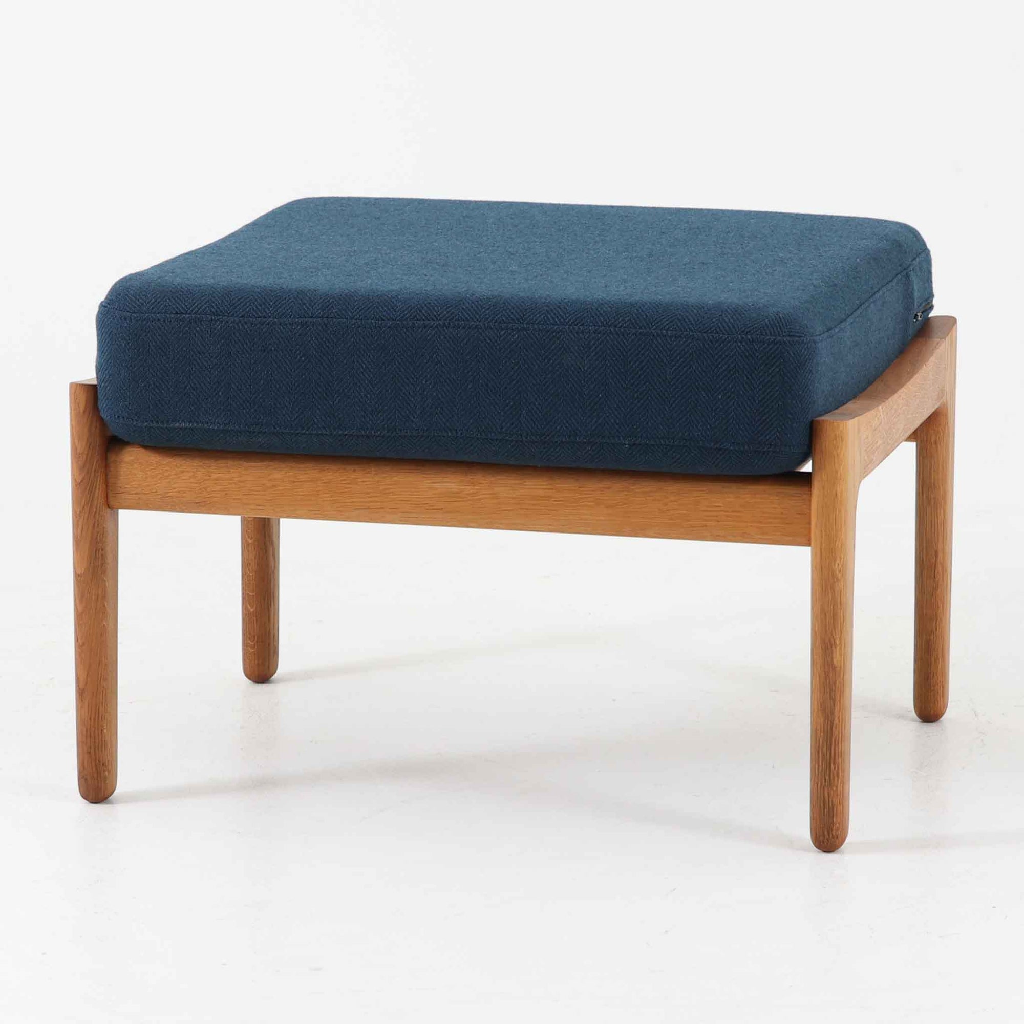 Hans J. WegnerがデザインしGetama社から発表されたGE290オットマンは、堅牢なオーク材のフレームと深みのあるブルーの張地が美しく調和した北欧ビンテージ家具です。直線的でありながら丸みを帯びた脚部が軽快な印象を与え、ソファと組み合わせてフットレストとしてはもちろん、単体でも腰掛けとして使用できます。経年による木肌の風合いと上質な張地が落ち着いた存在感を放つ逸品です。
