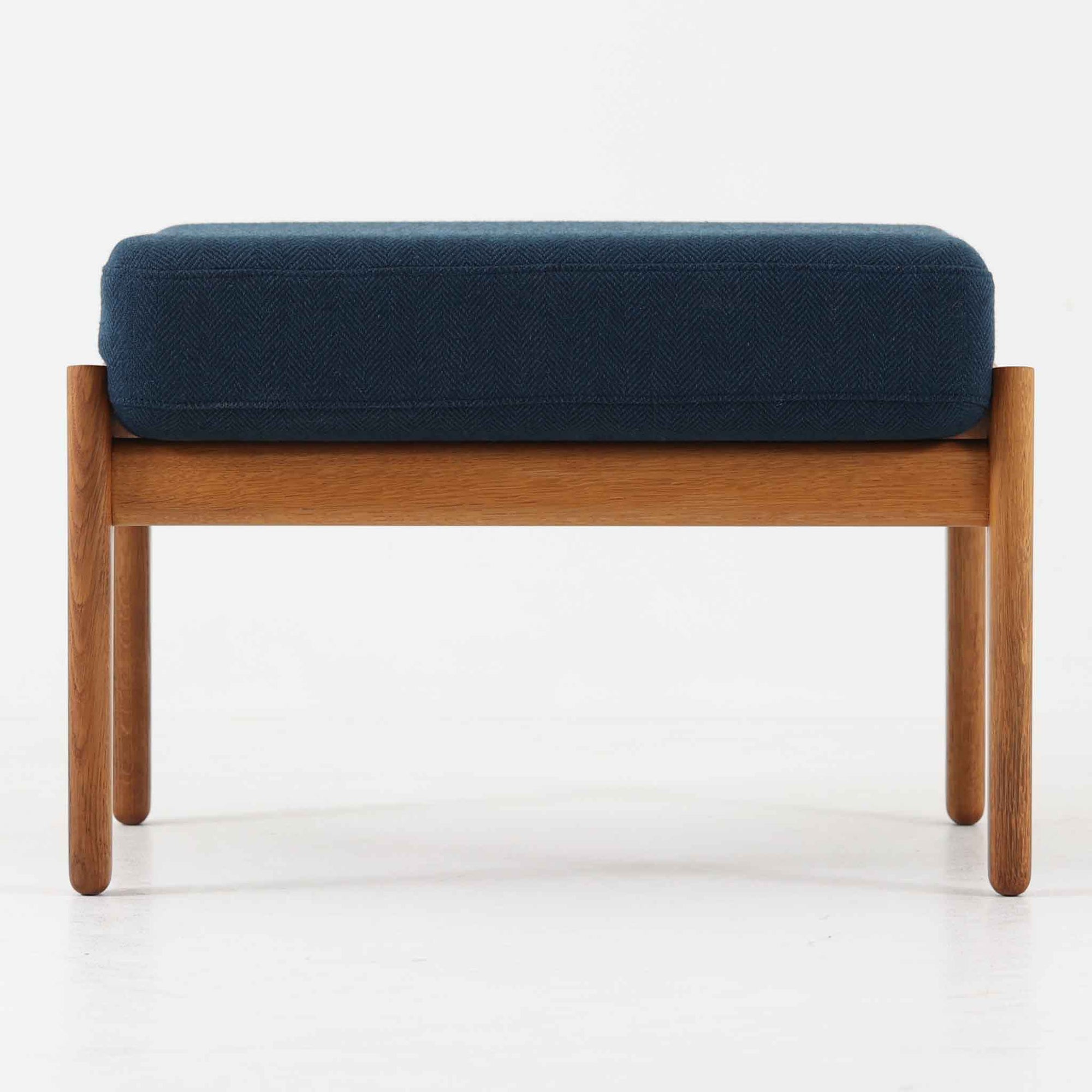 Hans J. WegnerがデザインしGetama社から発表されたGE290オットマンは、堅牢なオーク材のフレームと深みのあるブルーの張地が美しく調和した北欧ビンテージ家具です。直線的でありながら丸みを帯びた脚部が軽快な印象を与え、ソファと組み合わせてフットレストとしてはもちろん、単体でも腰掛けとして使用できます。経年による木肌の風合いと上質な張地が落ち着いた存在感を放つ逸品です。