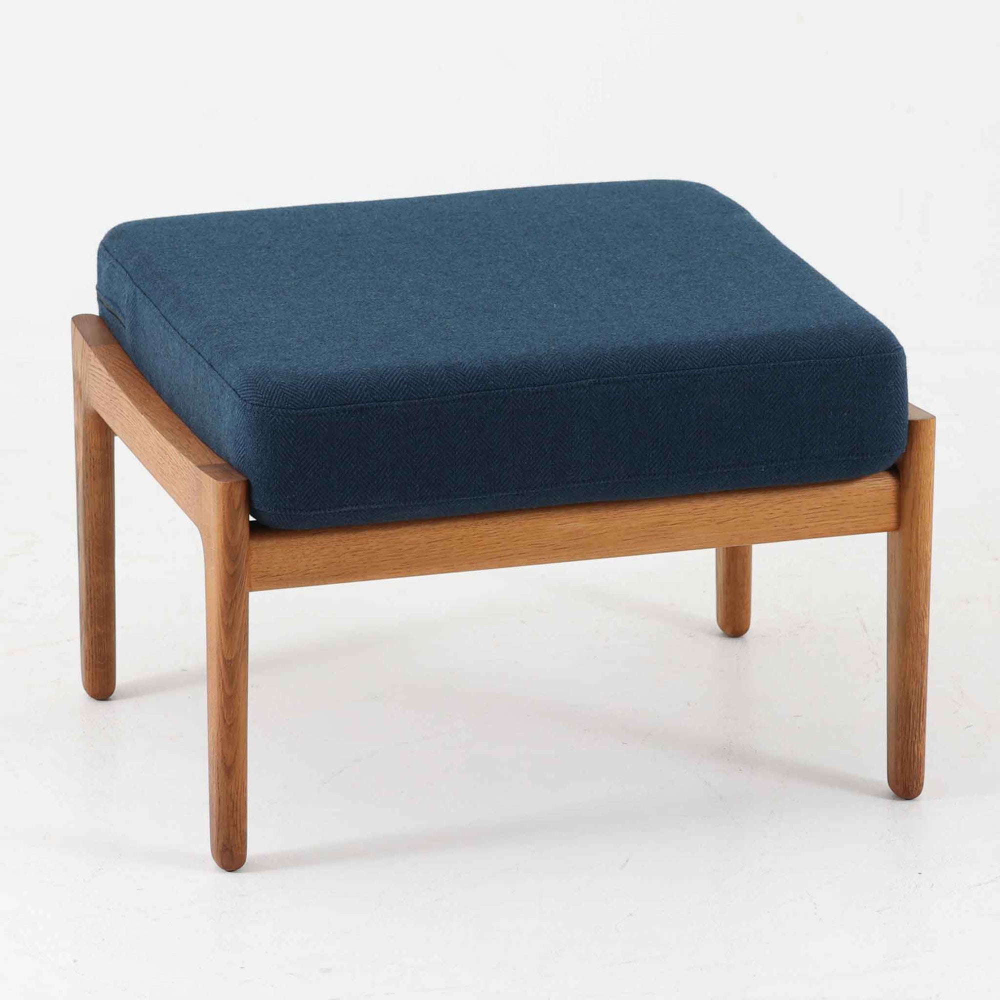 Hans J. WegnerがデザインしGetama社から発表されたGE290オットマンは、堅牢なオーク材のフレームと深みのあるブルーの張地が美しく調和した北欧ビンテージ家具です。直線的でありながら丸みを帯びた脚部が軽快な印象を与え、ソファと組み合わせてフットレストとしてはもちろん、単体でも腰掛けとして使用できます。経年による木肌の風合いと上質な張地が落ち着いた存在感を放つ逸品です。