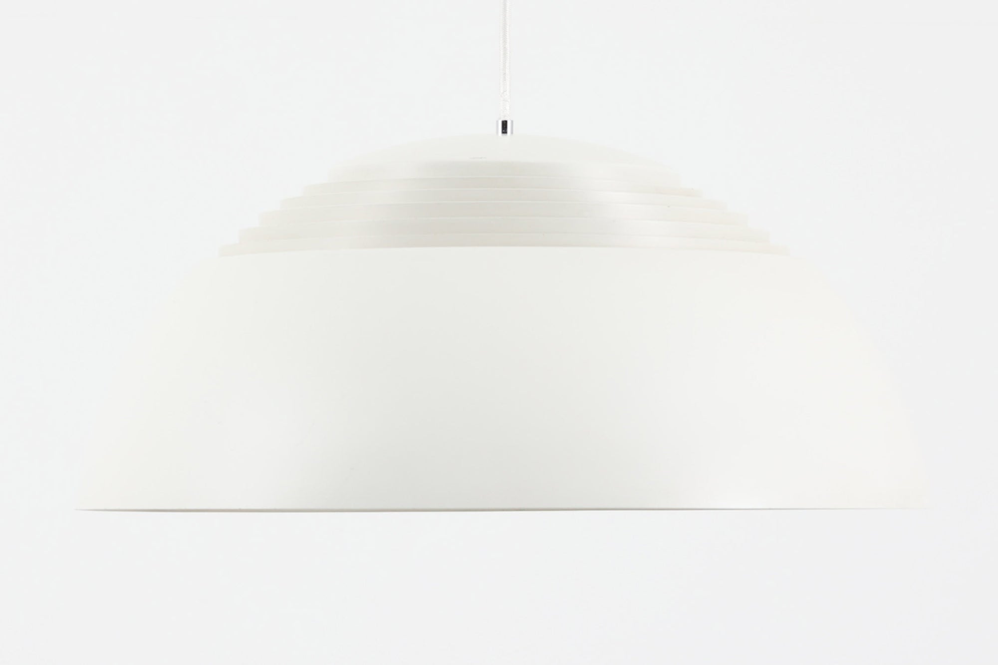 デンマークを代表する建築家・デザイナー、アルネ・ヤコブセンによってデザインされた「AJロイヤル」ペンダントライト。Louis Poulsen（ルイスポールセン）社より発表され、建築物と調和するミニマルなフォルムが特徴的です。こちらは直径50cmのホワイトカラーのモデルで、1958年に設計されたコペンハーゲンのSASロイヤルホテルのために開発されたシリーズのひとつ。上部のルーバー構造からも光が漏れる設計により、下方向だけでなく柔らかく空間全体を照らします。モダニズムの美学が息づく名作照明でありながら、日常の空間にもすっと馴染む柔らかな存在感が魅力です。ヴィンテージ品のため、シェードには擦り傷や小さな塗装剥がれ、内側に若干のサビが見られますが、使用には問題ありません。全体としては良好なコンディションを保っており、ヴィンテージならではの風合いをお楽しみいただけます。コードおよびソケットは国内基準に適合した仕様に交換済みで、すぐにご使用いただけます。