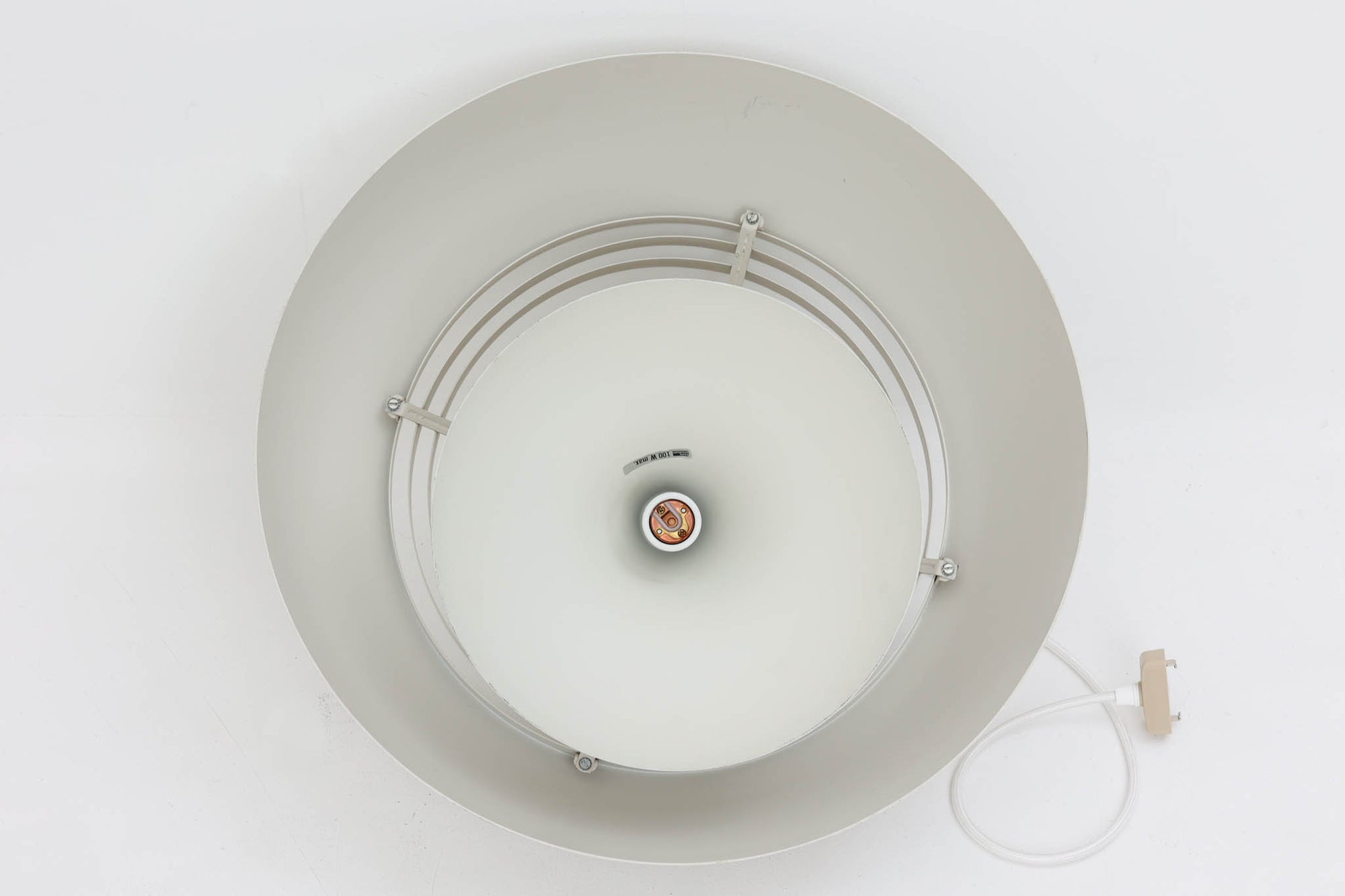 デンマークを代表する建築家・デザイナー、アルネ・ヤコブセンによってデザインされた「AJロイヤル」ペンダントライト。Louis Poulsen（ルイスポールセン）社より発表され、建築物と調和するミニマルなフォルムが特徴的です。こちらは直径50cmのホワイトカラーのモデルで、1958年に設計されたコペンハーゲンのSASロイヤルホテルのために開発されたシリーズのひとつ。上部のルーバー構造からも光が漏れる設計により、下方向だけでなく柔らかく空間全体を照らします。モダニズムの美学が息づく名作照明でありながら、日常の空間にもすっと馴染む柔らかな存在感が魅力です。ヴィンテージ品のため、シェードには擦り傷や小さな塗装剥がれ、内側に若干のサビが見られますが、使用には問題ありません。全体としては良好なコンディションを保っており、ヴィンテージならではの風合いをお楽しみいただけます。コードおよびソケットは国内基準に適合した仕様に交換済みで、すぐにご使用いただけます。