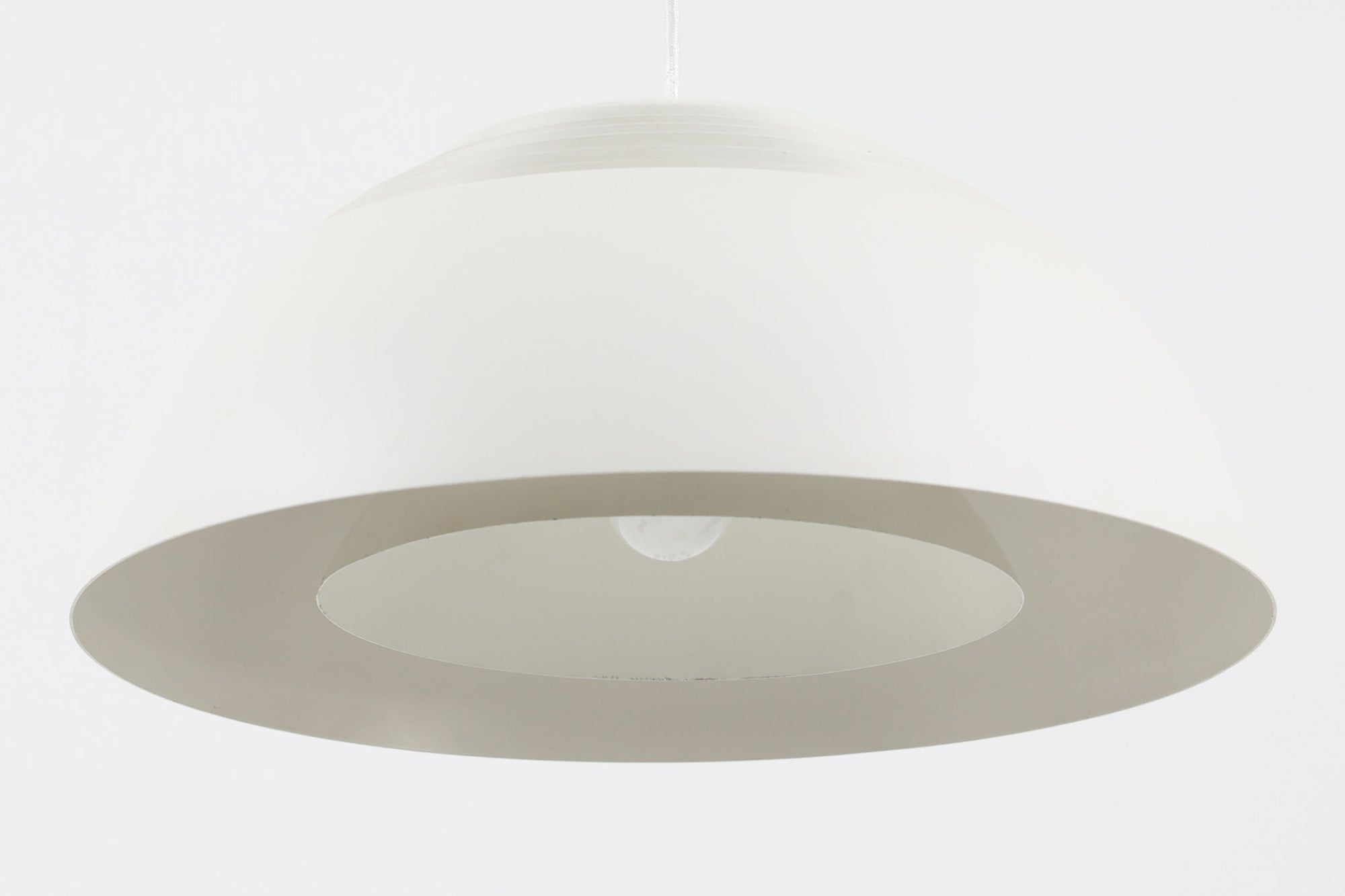 デンマークを代表する建築家・デザイナー、アルネ・ヤコブセンによってデザインされた「AJロイヤル」ペンダントライト。Louis Poulsen（ルイスポールセン）社より発表され、建築物と調和するミニマルなフォルムが特徴的です。こちらは直径50cmのホワイトカラーのモデルで、1958年に設計されたコペンハーゲンのSASロイヤルホテルのために開発されたシリーズのひとつ。上部のルーバー構造からも光が漏れる設計により、下方向だけでなく柔らかく空間全体を照らします。モダニズムの美学が息づく名作照明でありながら、日常の空間にもすっと馴染む柔らかな存在感が魅力です。ヴィンテージ品のため、シェードには擦り傷や小さな塗装剥がれ、内側に若干のサビが見られますが、使用には問題ありません。全体としては良好なコンディションを保っており、ヴィンテージならではの風合いをお楽しみいただけます。コードおよびソケットは国内基準に適合した仕様に交換済みで、すぐにご使用いただけます。