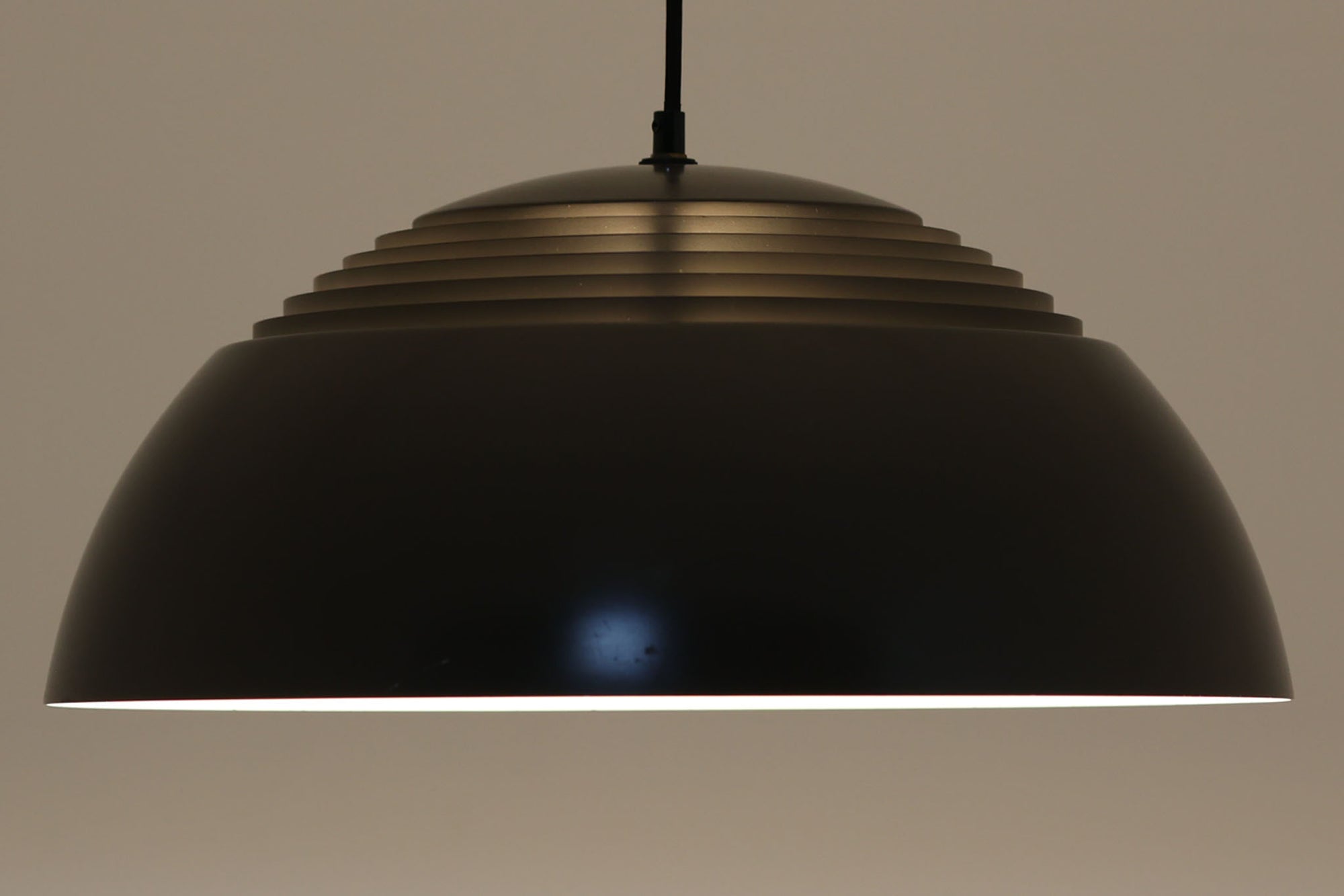 アルネ・ヤコブセンによってデザインされ、Louis Poulsen（ルイスポールセン）より製造されたAJロイヤル ペンダントライト。こちらは希少なグラファイトグレーカラーで、落ち着いたトーンが空間にシックな雰囲気を演出します。直径50cmの大型サイズで、光源を遮る構造と放射状に配置されたルーバーにより、眩しさを抑えた柔らかい光が空間全体を包み込みます。点灯時には上部のスリットから漏れる光も美しく、デザイン性と機能性を兼ね備えた一品です。外観には小傷や使用感がありますが、ビンテージならではの風合いとしてお楽しみいただけます。