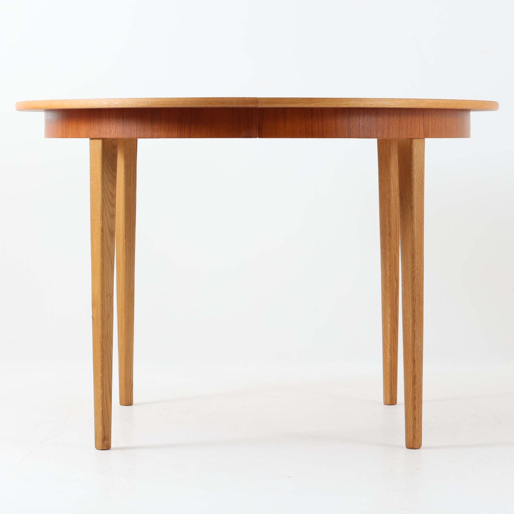 チーク材の天板とオーク材の脚部が美しく調和したこちらの円形エクステンションダイニングテーブルは、デンマーク製ならではの丁寧な仕上げと実用性が魅力の北欧ビンテージ家具です。直径110cmの円形天板は、中央に拡張板を2枚追加することで広々としたオーバル型に変化し、来客時やホームパーティーなどにも柔軟に対応できます。細身のテーパー脚が軽やかな印象を与え、温かみのある木の表情が空間に自然と溶け込む逸品です。