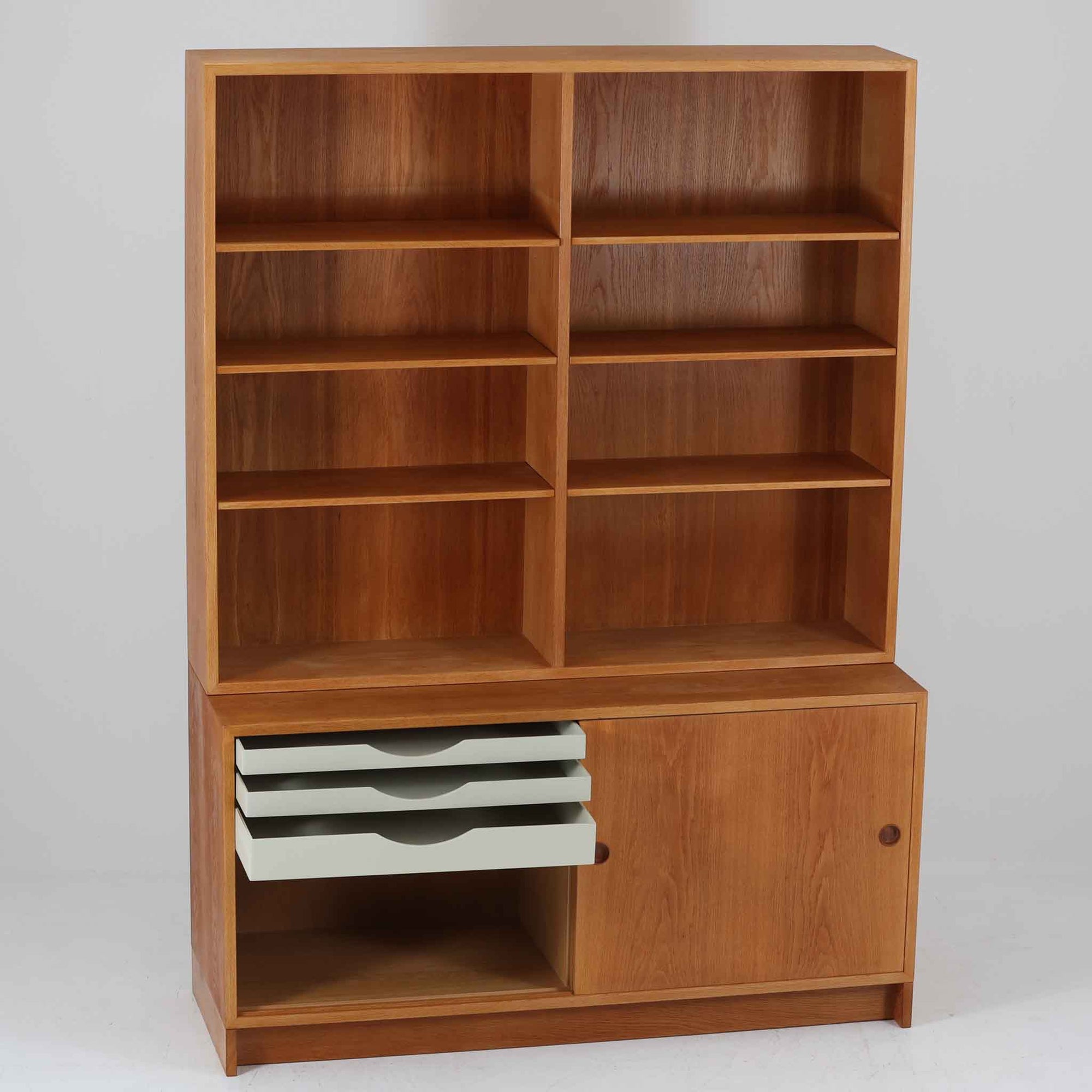 スウェーデンのKarl Andersson社より、Borge Mogensenによってデザインされたディスプレイシェルフは、端正な佇まいと機能性を兼ね備えた北欧ビンテージ家具です。オーク材の温もりある質感と美しい木目が魅力で、上段は可動棚付きのオープンシェルフ、下段には丸い掘り込み取手がアクセントとなった引き戸収納が配されています。文庫本や器、日用品の整理に最適で、暮らしの中で永く使い続けられる名作です。