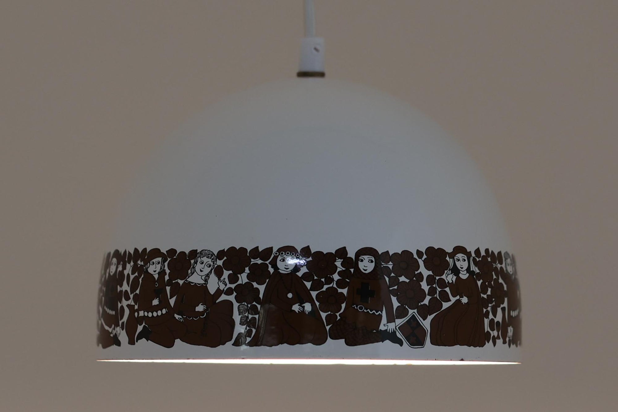 フィンランドの名窯ARABIA（アラビア）社によるRITARI（リタリ）シリーズのビンテージランプシェードです。柔らかなカーブを描くドーム型のフォルムは、フィンランドモダンを代表するデザイナーKaj Franck（カイ・フランク）によるもので、花と人物が描かれた印象的な装飾はRaija Uosikkinen（ライヤ・ウオシッキネン）によってデザインされました。RITARIはフィンランド語で「騎士」を意味し、中世の衣装をまとった人物たちが、ブラウンを基調とした花々とともにシェードをぐるりと囲むユニークでノスタルジックなデザインが特徴です。素材はホーロー（琺瑯）製で、しっかりとした重量感と経年による独特の風合いも楽しめるアイテム。点灯時にはやわらかな光を下方向に届け、空間に温もりを添えてくれます。北欧らしいストーリー性とデザイン性を併せ持った、希少なビンテージ照明をお探しの方におすすめです。