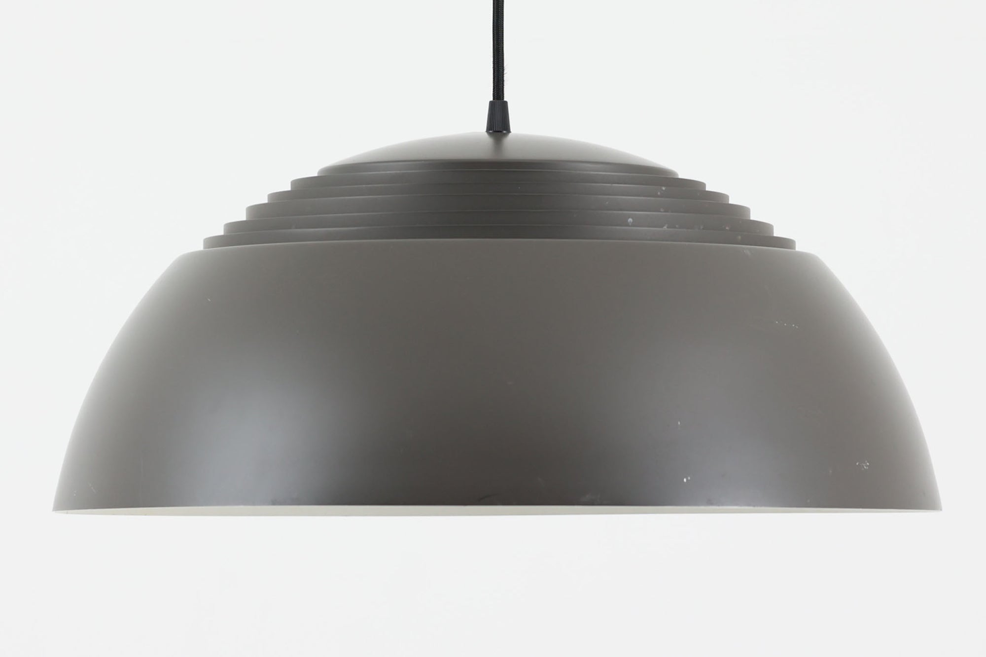 アルネ・ヤコブセンによってデザインされ、Louis Poulsen（ルイスポールセン）より製造されたAJロイヤル ペンダントライト。こちらは希少なグラファイトグレーカラーで、落ち着いたトーンが空間にシックな雰囲気を演出します。直径50cmの大型サイズで、光源を遮る構造と放射状に配置されたルーバーにより、眩しさを抑えた柔らかい光が空間全体を包み込みます。点灯時には上部のスリットから漏れる光も美しく、デザイン性と機能性を兼ね備えた一品です。外観には小傷や使用感がありますが、ビンテージならではの風合いとしてお楽しみいただけます。