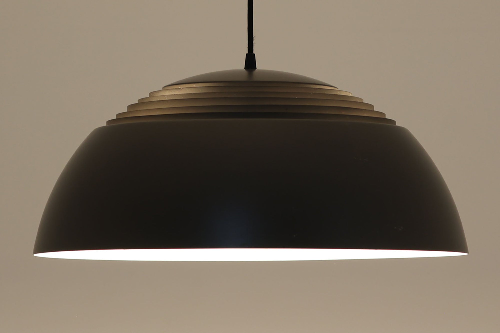 アルネ・ヤコブセンによってデザインされ、Louis Poulsen（ルイスポールセン）より製造されたAJロイヤル ペンダントライト。こちらは希少なグラファイトグレーカラーで、落ち着いたトーンが空間にシックな雰囲気を演出します。直径50cmの大型サイズで、光源を遮る構造と放射状に配置されたルーバーにより、眩しさを抑えた柔らかい光が空間全体を包み込みます。点灯時には上部のスリットから漏れる光も美しく、デザイン性と機能性を兼ね備えた一品です。外観には小傷や使用感がありますが、ビンテージならではの風合いとしてお楽しみいただけます。