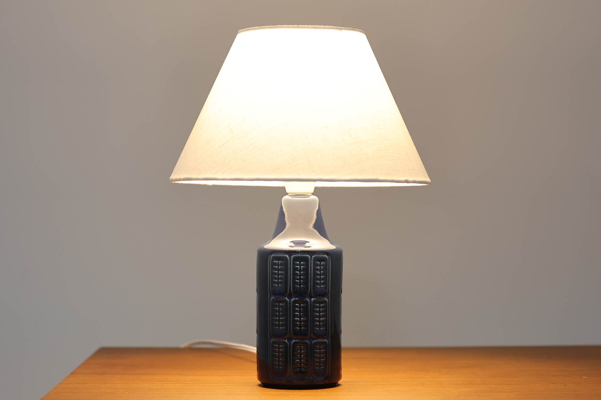 デンマークの陶器ブランドDesiree（デシレ）によって制作されたヴィンテージのテーブルランプ。深みのあるネイビーブルーの艶やかな釉薬に、規則正しく並んだ凹凸のある幾何学模様が印象的なデザインです。スリムな円筒形のフォルムは空間にすっきりと馴染みつつ、どこかグラフィカルな存在感を漂わせます。底部には「Desiree Stentoj – Denmark」の刻印と品番が施されています。シェードには新品のリネン製シェードを使用し、光をやわらかく透過。北欧ヴィンテージらしいモダンさと温もりを兼ね備えた一灯です。