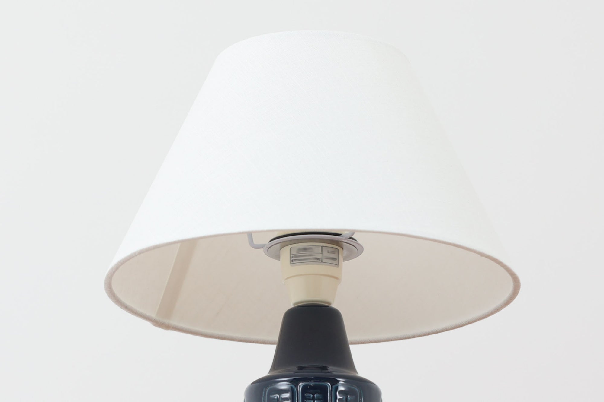 デンマークの陶器ブランドDesiree（デシレ）によって制作されたヴィンテージのテーブルランプ。深みのあるネイビーブルーの艶やかな釉薬に、規則正しく並んだ凹凸のある幾何学模様が印象的なデザインです。スリムな円筒形のフォルムは空間にすっきりと馴染みつつ、どこかグラフィカルな存在感を漂わせます。底部には「Desiree Stentoj – Denmark」の刻印と品番が施されています。シェードには新品のリネン製シェードを使用し、光をやわらかく透過。北欧ヴィンテージらしいモダンさと温もりを兼ね備えた一灯です。