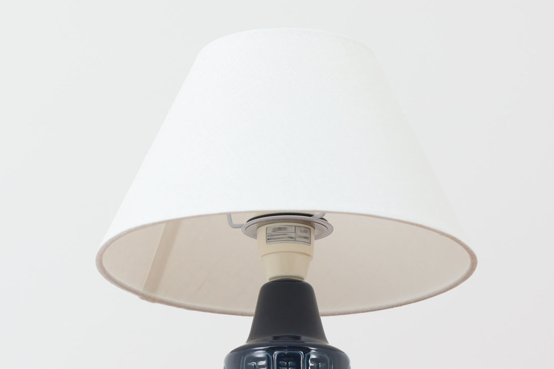 デンマークの陶器ブランドDesiree（デシレ）によって制作されたヴィンテージのテーブルランプ。深みのあるネイビーブルーの艶やかな釉薬に、規則正しく並んだ凹凸のある幾何学模様が印象的なデザインです。スリムな円筒形のフォルムは空間にすっきりと馴染みつつ、どこかグラフィカルな存在感を漂わせます。底部には「Desiree Stentoj – Denmark」の刻印と品番が施されています。シェードには新品のリネン製シェードを使用し、光をやわらかく透過。北欧ヴィンテージらしいモダンさと温もりを兼ね備えた一灯です。