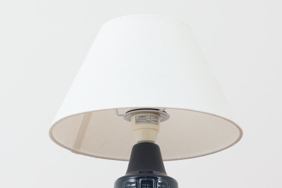 デンマークの陶器ブランドDesiree（デシレ）によって制作されたヴィンテージのテーブルランプ。深みのあるネイビーブルーの艶やかな釉薬に、規則正しく並んだ凹凸のある幾何学模様が印象的なデザインです。スリムな円筒形のフォルムは空間にすっきりと馴染みつつ、どこかグラフィカルな存在感を漂わせます。底部には「Desiree Stentoj – Denmark」の刻印と品番が施されています。シェードには新品のリネン製シェードを使用し、光をやわらかく透過。北欧ヴィンテージらしいモダンさと温もりを兼ね備えた一灯です。
