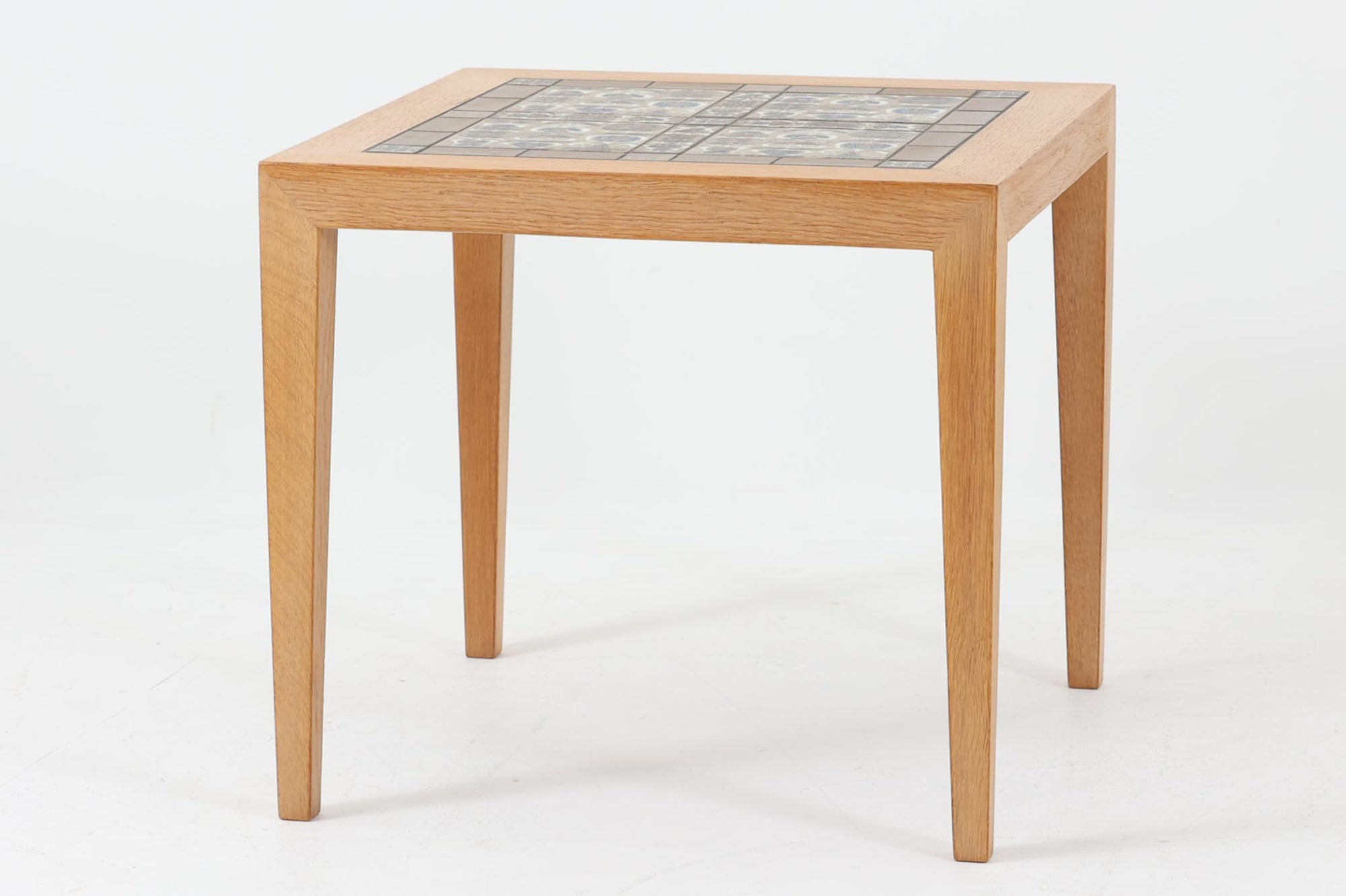 デンマークの家具デザイナー、Severin Hansen Jr.によってデザインされたビンテージのサイドテーブルです。製造はデンマークの名門家具メーカー「Haslev（ハスレヴ）」社。天板にはRoyal Copenhagen（ロイヤルコペンハーゲン）のアートタイルが贅沢にあしらわれています。タイルはNils Thorsson（ニルス・トーソン）が手掛けた人気の「BACA（バッカ）」シリーズ。象徴的なブルーとブラウンの色使いと、有機的かつ幾何学的なモチーフが印象的で、アートピースとしての存在感を放ちます。テーブル本体はオーク材を使用しており、Severin Hansen Jr.の特徴でもあるシャープなカットが施されたコーナーと、すっきりとしたテーパードレッグが、シンプルながら洗練された美しさを醸し出しています。ヴィンテージならではの深い味わいと、アート性・機能性を兼ね備えた一台。リビングやベッドサイドに、さりげなく上質な北欧デザインを取り入れてみてはいかがでしょうか。