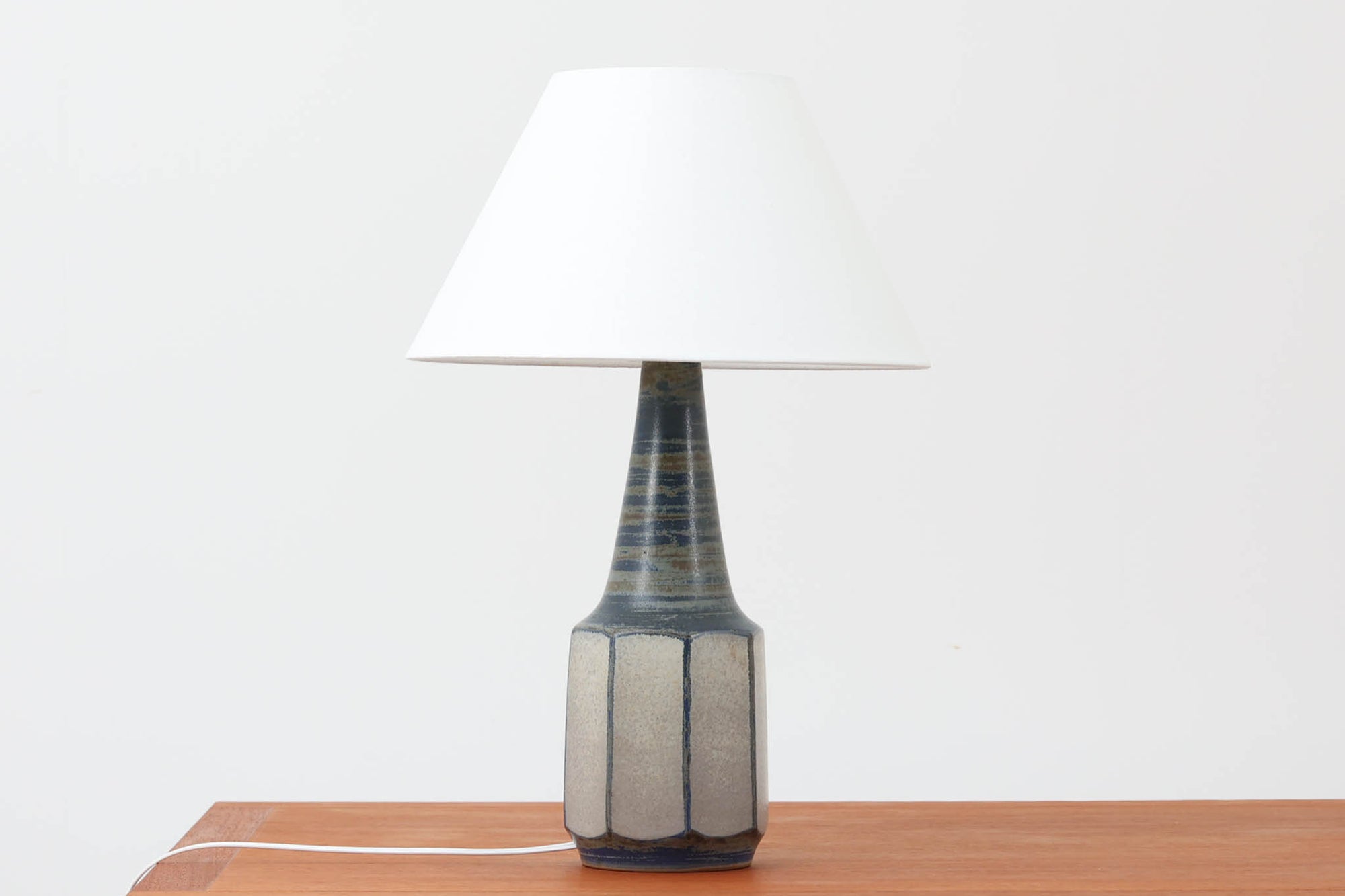 デンマーク・ボーンホルム島の名窯michael Andersen & Sonによるテーブルランプ。八角形のシャープなフォルムと、マットな質感の釉薬のコントラストが美しい一点です。落ち着いたブルーグレーのトーンがインテリアに静かなアクセントを添えてくれます。高さは約50cmとやや大きめで、程よい存在感があり、サイドボードやコンソールの上などにもおすすめです。シェードは新品のリネン製。