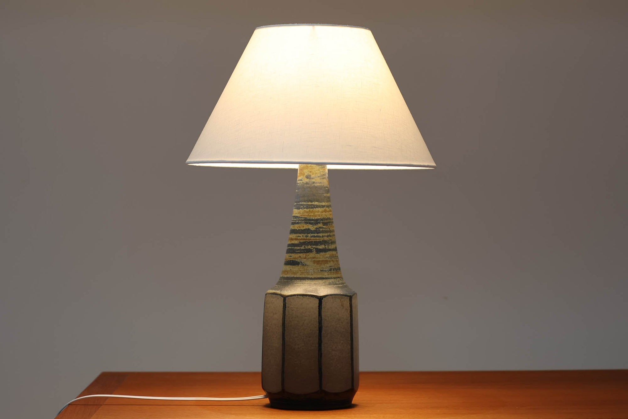 デンマーク・ボーンホルム島の名窯michael Andersen & Sonによるテーブルランプ。八角形のシャープなフォルムと、マットな質感の釉薬のコントラストが美しい一点です。落ち着いたブルーグレーのトーンがインテリアに静かなアクセントを添えてくれます。高さは約50cmとやや大きめで、程よい存在感があり、サイドボードやコンソールの上などにもおすすめです。シェードは新品のリネン製。