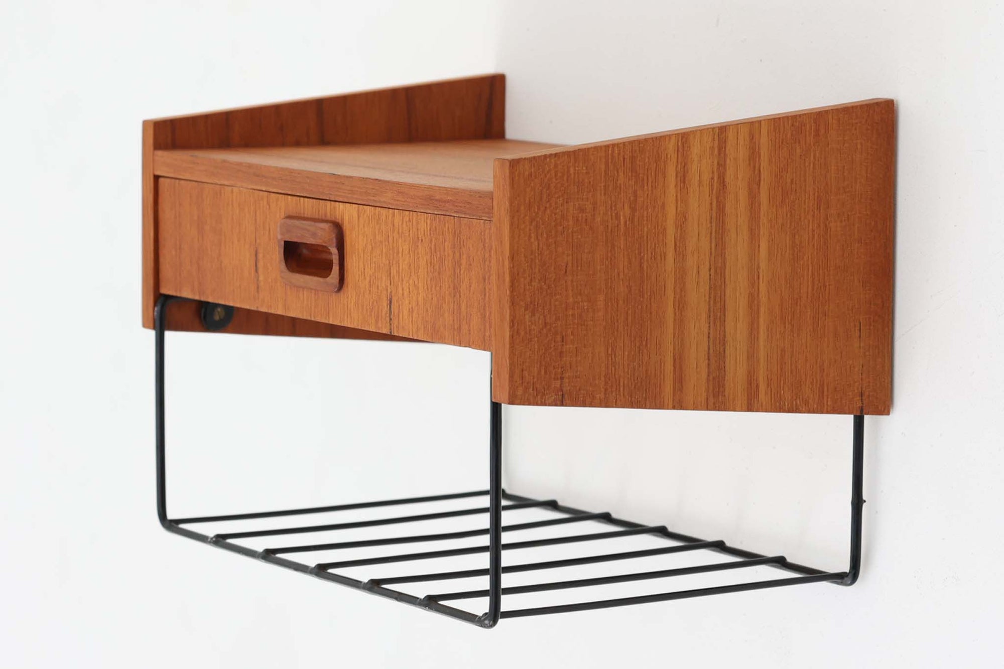 スウェーデン製のヴィンテージウォールシェルフ。温かみのあるチーク材と、ブラックスチールのコンビネーションが魅力的な一台です。シンプルな構成ながら、前面の丸みを帯びた掘り込み取手や、緩やかな角度のついたサイドパネルなど、北欧デザインらしい細やかなディテールが随所に見られます。引き出しには小物類をすっきりと収納でき、下部のスチールラックはマフラーや雑誌、スリッパなどの置き場としても便利。玄関やリビング、寝室など、さまざまな空間にフィットする汎用性の高いアイテムです。