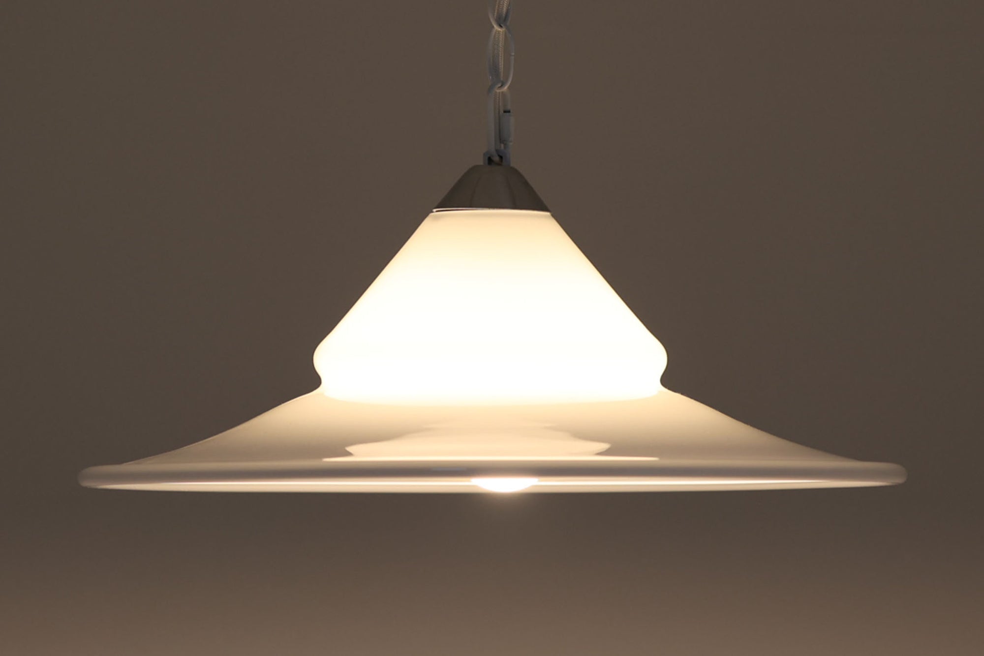 デンマークの名窯、Royal Copenhagen（ロイヤルコペンハーゲン）によるビンテージペンダントライト「Opus」。直径44cmのガラスシェードは、乳白色の滑らかな質感と優美な曲線を持ち、点灯時には柔らかな光をふんわりと拡散します。シェードの中央部にさりげない段差を設けたデザインは、シンプルながらも立体感があり、空間に洗練されたアクセントを加えます。金属パーツにはやや経年の使用感が見られますが、シェード自体は非常に良好な状態で、ガラスの透明感と清潔感を十分に感じられます。ダイニングやリビング、吹き抜けの空間など、幅広いインテリアにマッチし、北欧モダンな空間演出にぴったりの一灯です。
