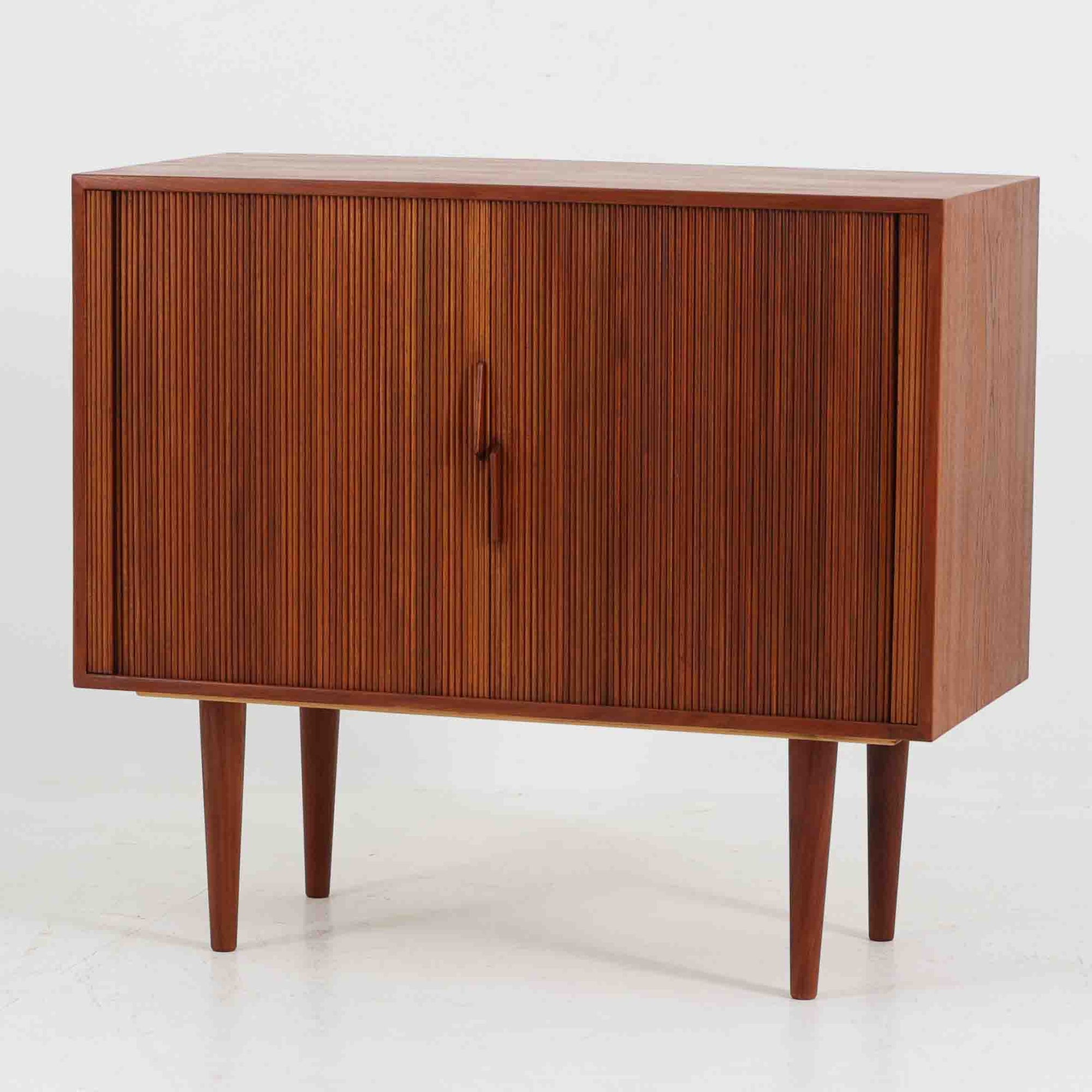 Kai Kristiansen（カイ・クリスチャンセン）によるデザインで、Feldballes Mobelfabrik社から製造された蛇腹キャビネットです。繊細なスリットが並ぶチーク材のロールフロントは職人技が光る美しい仕上がりで、滑らかな開閉動作も魅力のひとつ。内部は仕切り板と小収納が備えられ、実用性にも優れています。右下の小収納部は前板のみが貼られた飾り構造で、内部に収納はありません。特注仕様と思われる希少な一点です。シンプルながら存在感があり、リビングやワークスペースにも調和する北欧ビンテージらしい端正な佇まいです。
