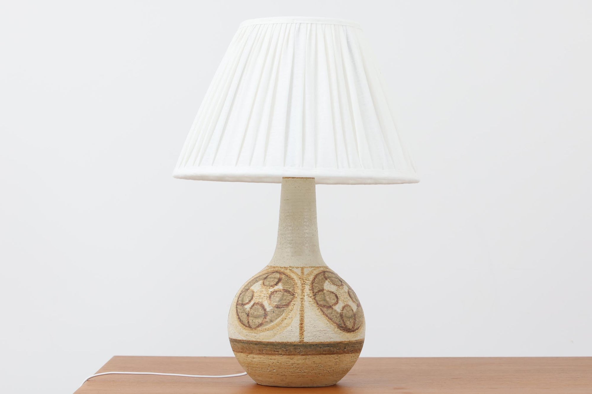 デンマーク・ボーンホルム島の名窯 SOHOLM（スーホルム）によるヴィンテージのテーブルランプです。ふっくらとしたボディに施された幾何学模様が印象的で、素朴であたたかみのある釉薬の風合いが魅力。北欧らしいナチュラルな空気感を空間に添えてくれます。高さは約48cmとやや大きめで、しっかりとした存在感がありながらも落ち着いた佇まいです。シェードは新品のリネン製に交換済み。やさしい光がファブリック越しにふんわりと広がり、ベッドサイドやリビングのアクセントにぴったりです。北欧陶器ならではの土の質感と落ち着いた色彩をお楽しみください。